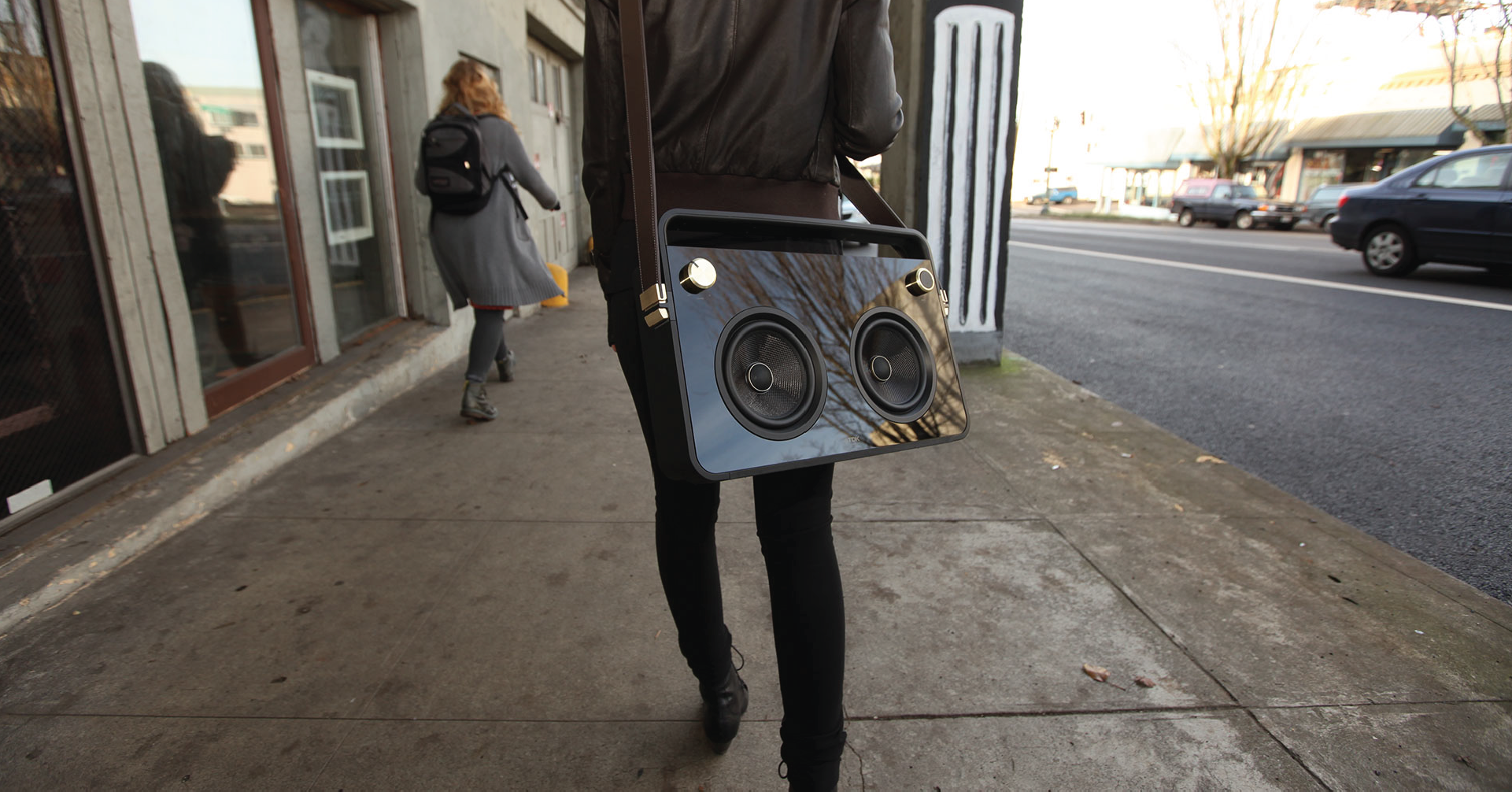 Person walking on a sidewalk carrying a black boombox with gold knobs and a brown leather strap