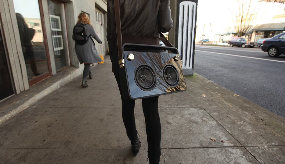 Person walking on a sidewalk carrying a black boombox with gold knobs and a brown leather strap