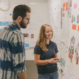 Two people smiling and looking at a whiteboard with sticky notes on it