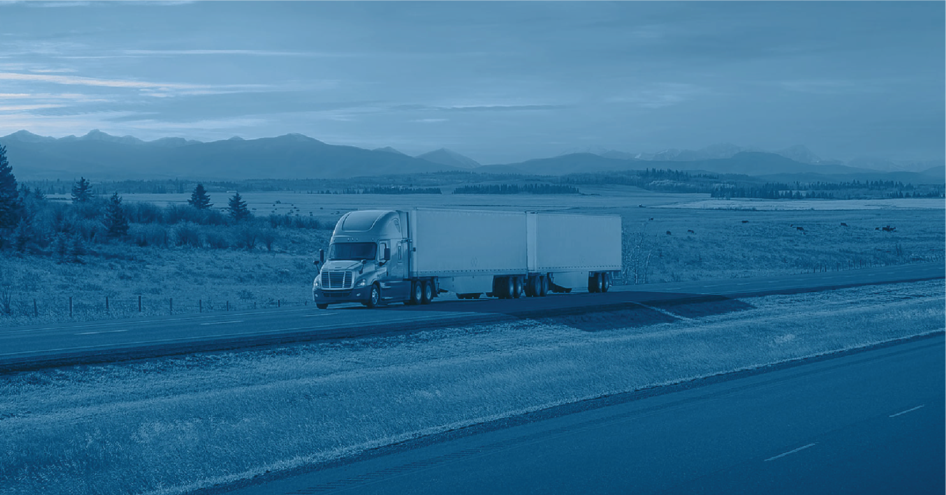 Semi truck driving on a highway with a field, trees, and a mountain range in the background