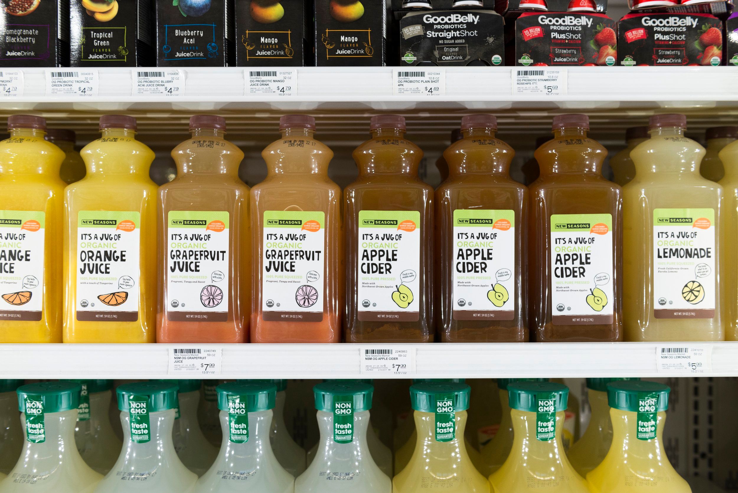 A row of juices made by New Seasons including orange juice, grapefruit juice, and apple cider, placed in a refrigerator next to other brands of juice