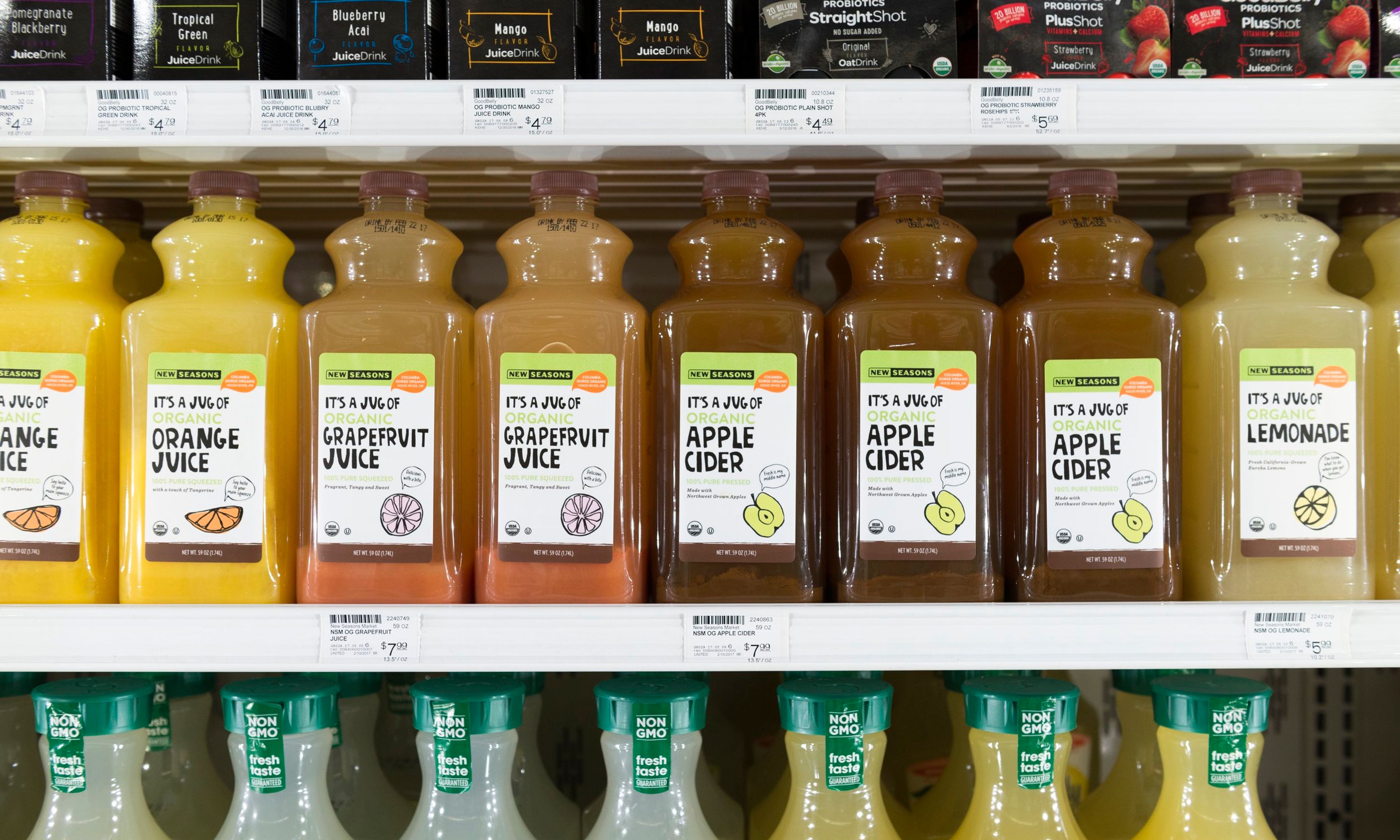 A row of juices made by New Seasons including orange juice, grapefruit juice, and apple cider, placed in a refrigerator next to other brands of juice