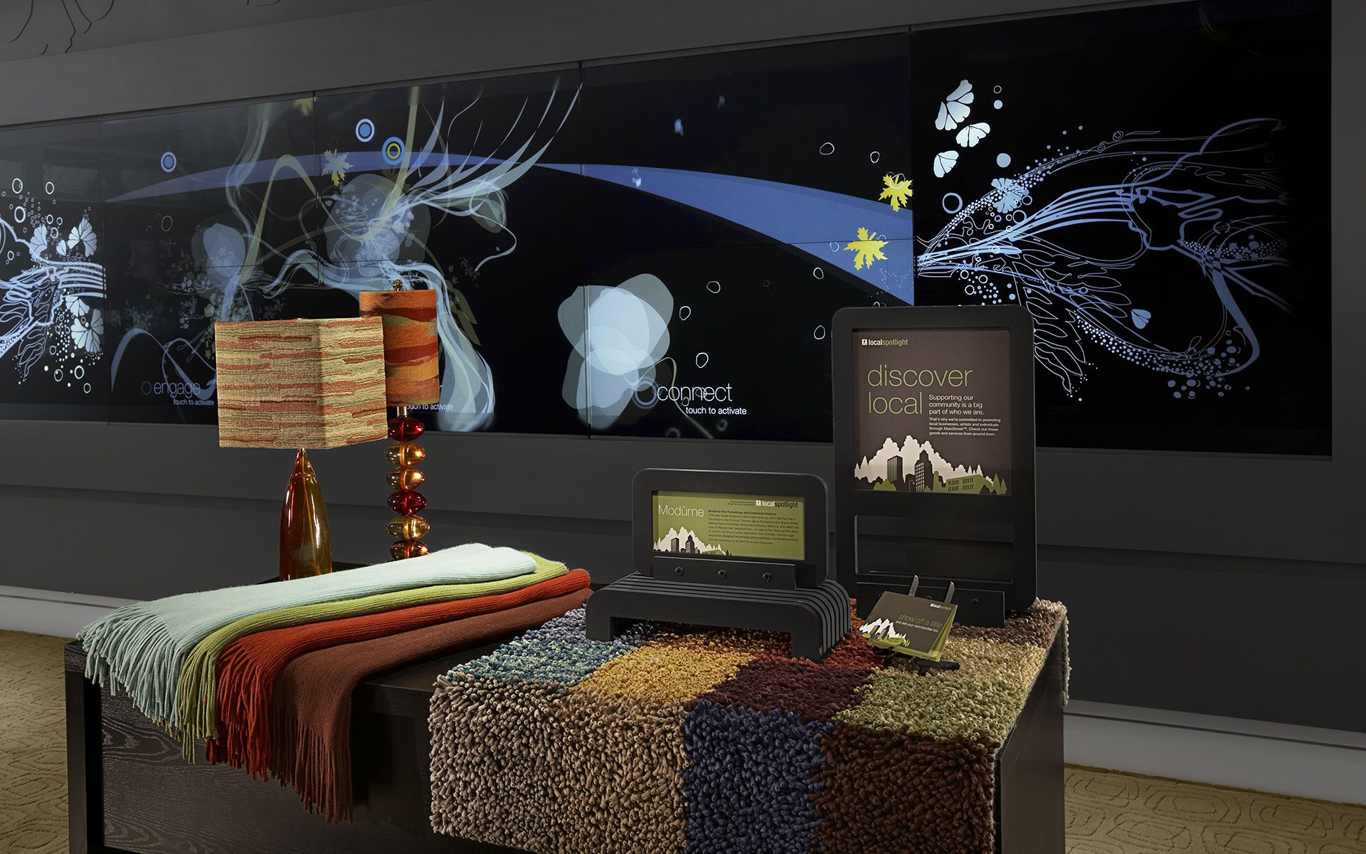 Tables with colorful rugs and lamps in front of a digital display showing animated illustrations on a touch screen