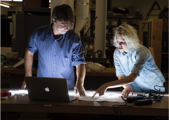 Two people pointing at an illuminated surface in front of an open laptop