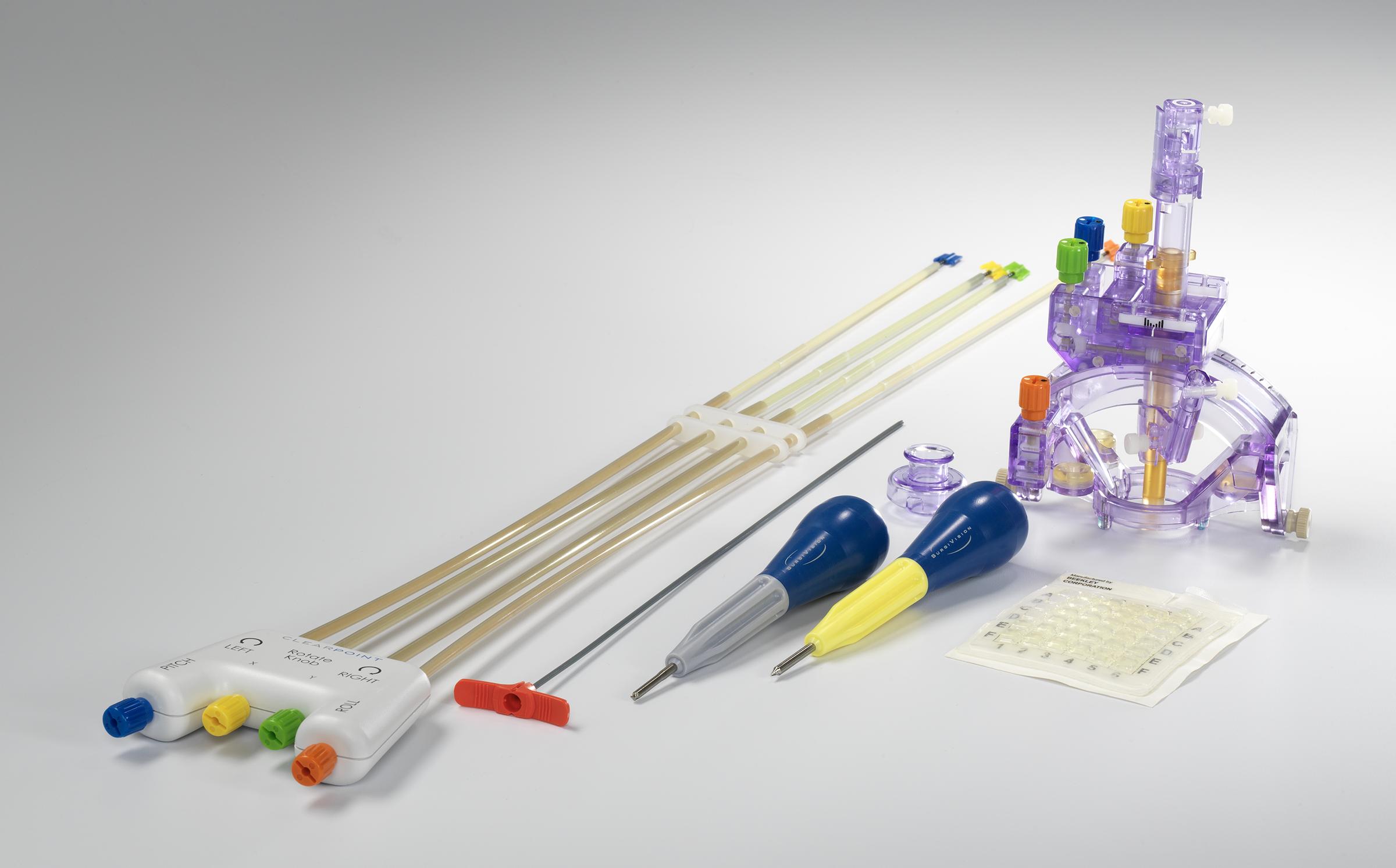 Various components of a surgical device, including four rubber tubes attached to a white handle, a purple plastic component that has four colored knobs attached to the top and side, a long metal needle with an orange winged handle on the top, and two specialized screwdrivers