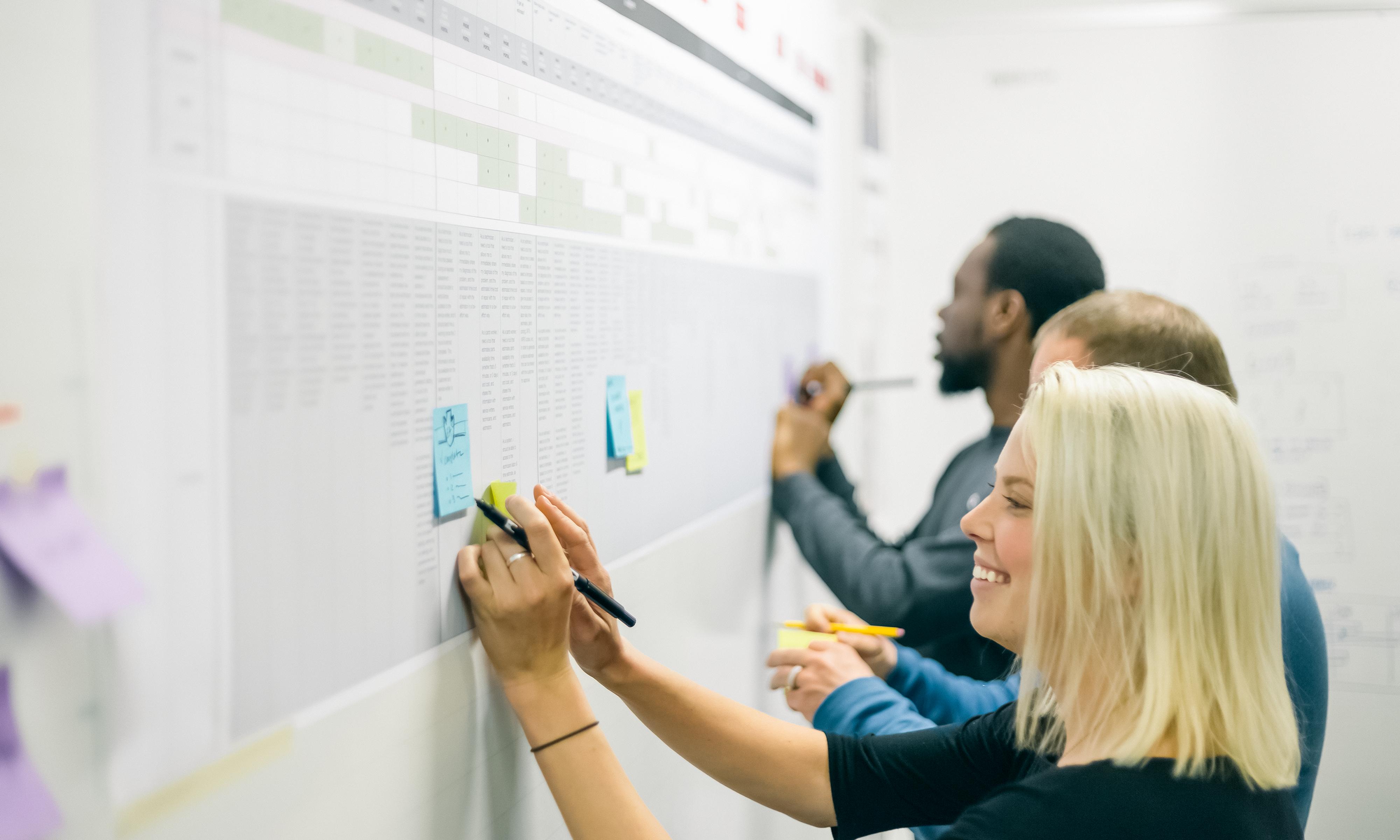 Three people writing on sticky notes that are placed on a whiteboard