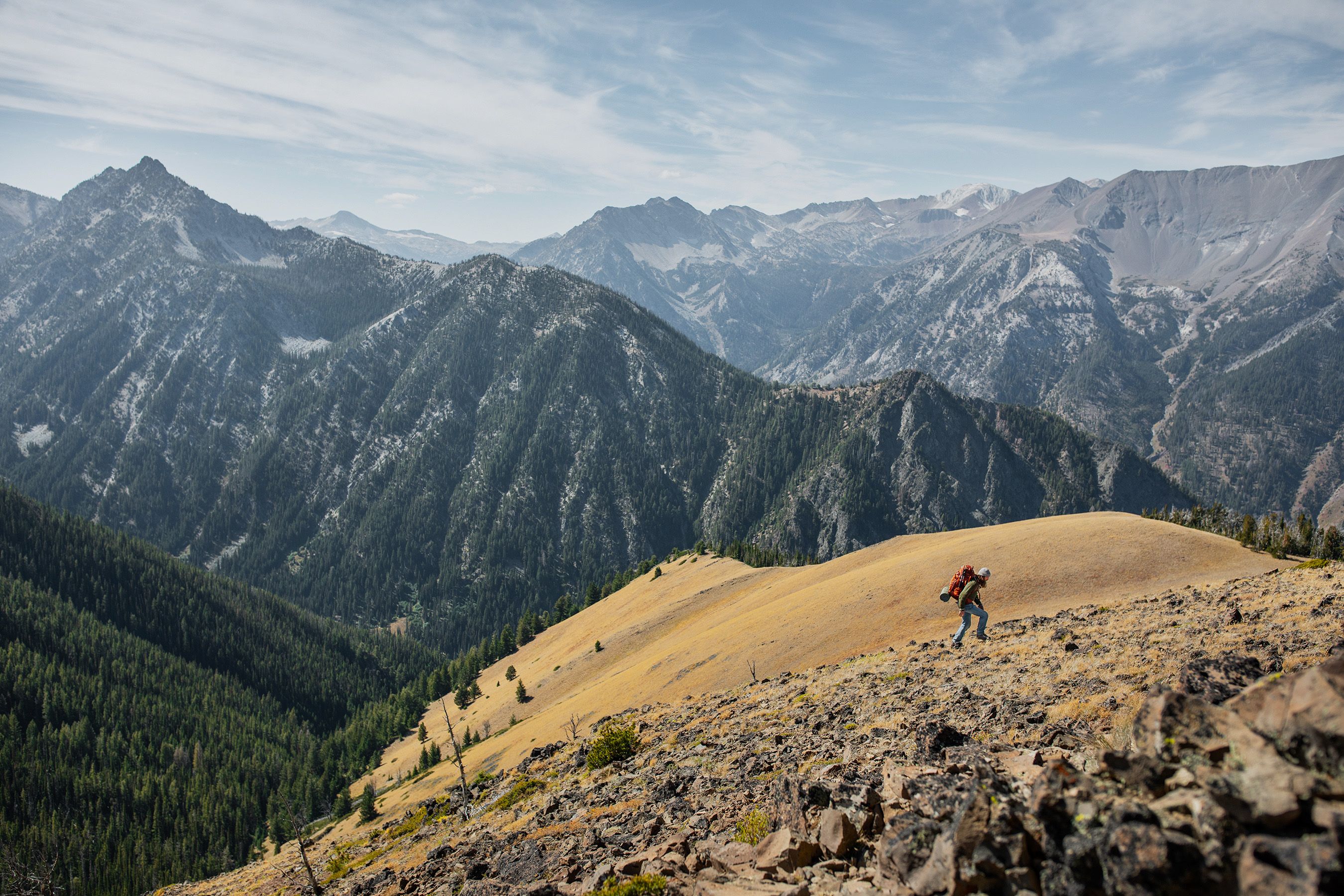 Person wearing a large backpack hiking up a mountain side in front of a mountain range covered in trees