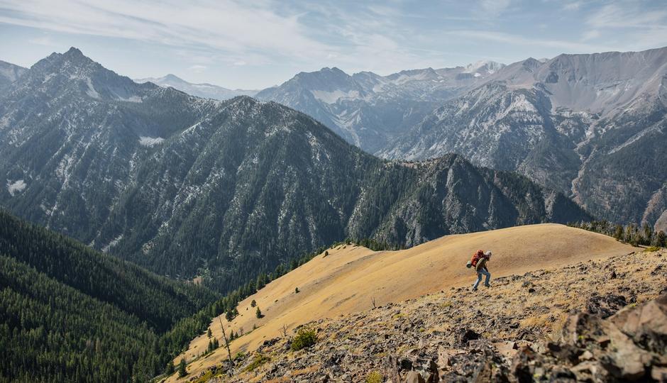 Person wearing a large backpack hiking up a mountain side in front of a mountain range covered in trees