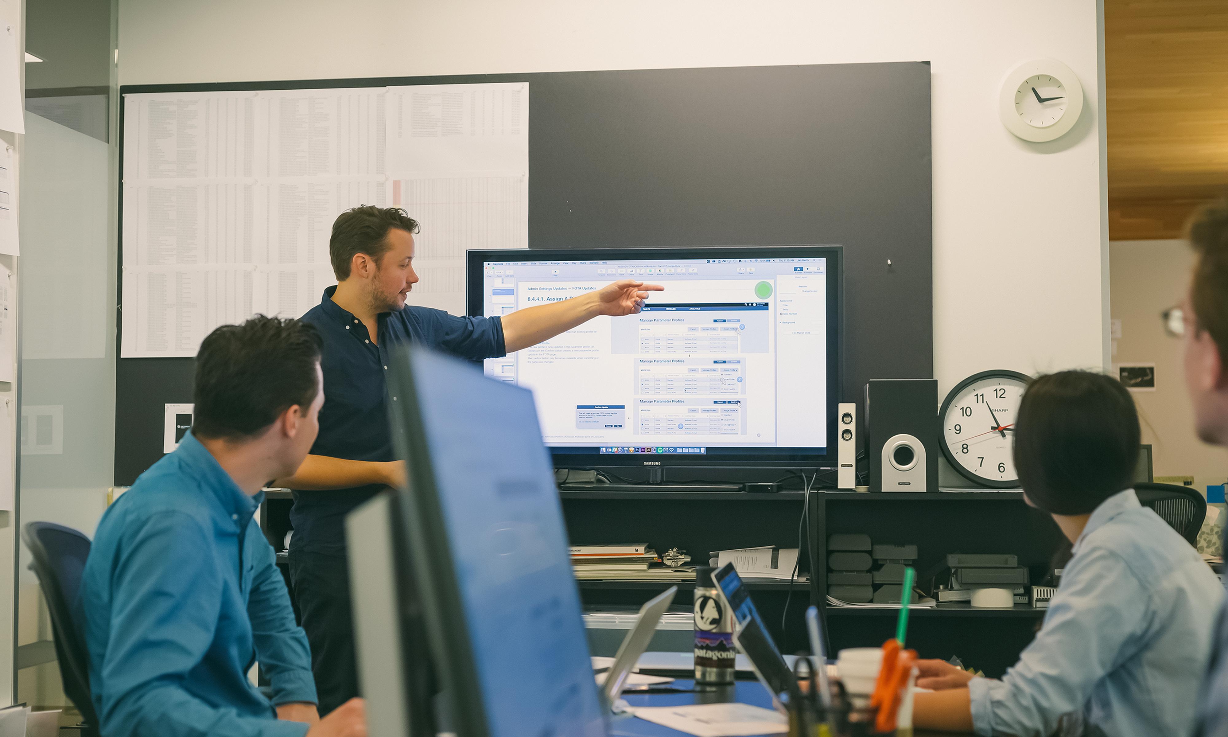 Three people sitting at a conference table, looking at one person who is pointing to a monitor that is displaying several data tables