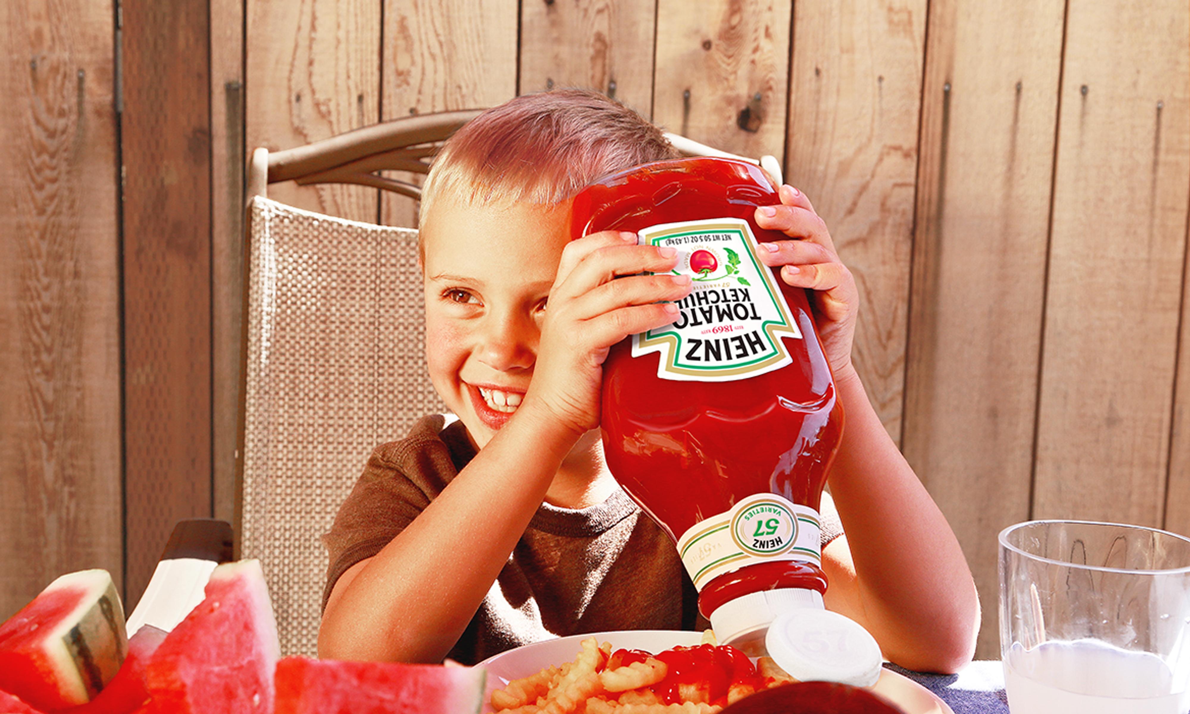 Child smiling and holding a Heinz ketchup bottle upside down above a plate of fries with ketchup on them