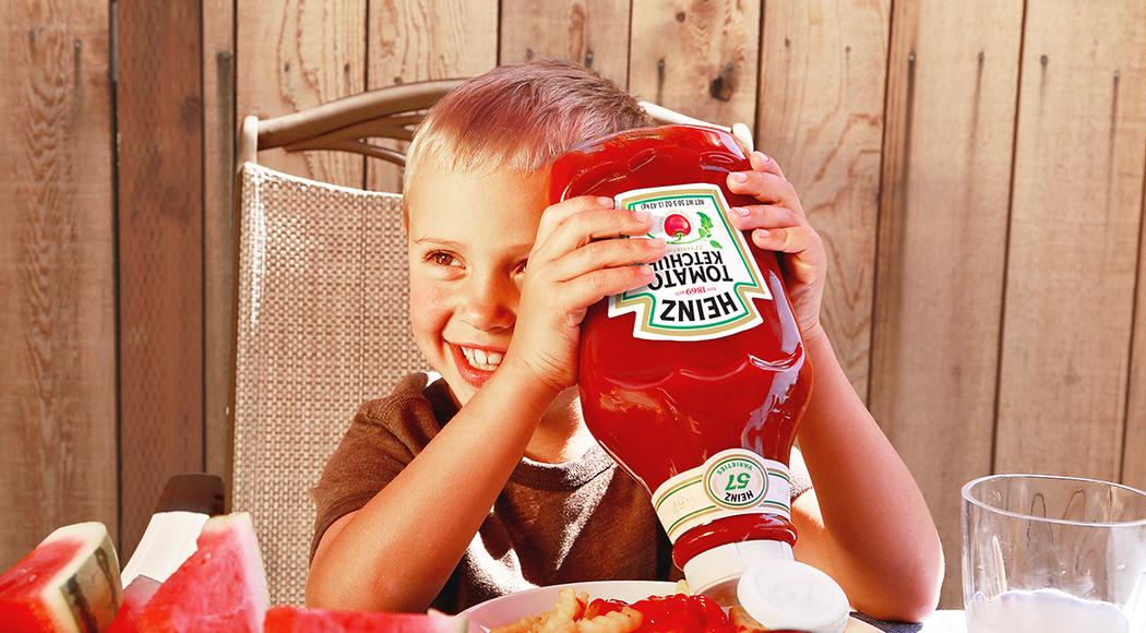 Child smiling and holding a Heinz ketchup bottle upside down above a plate of fries with ketchup on them