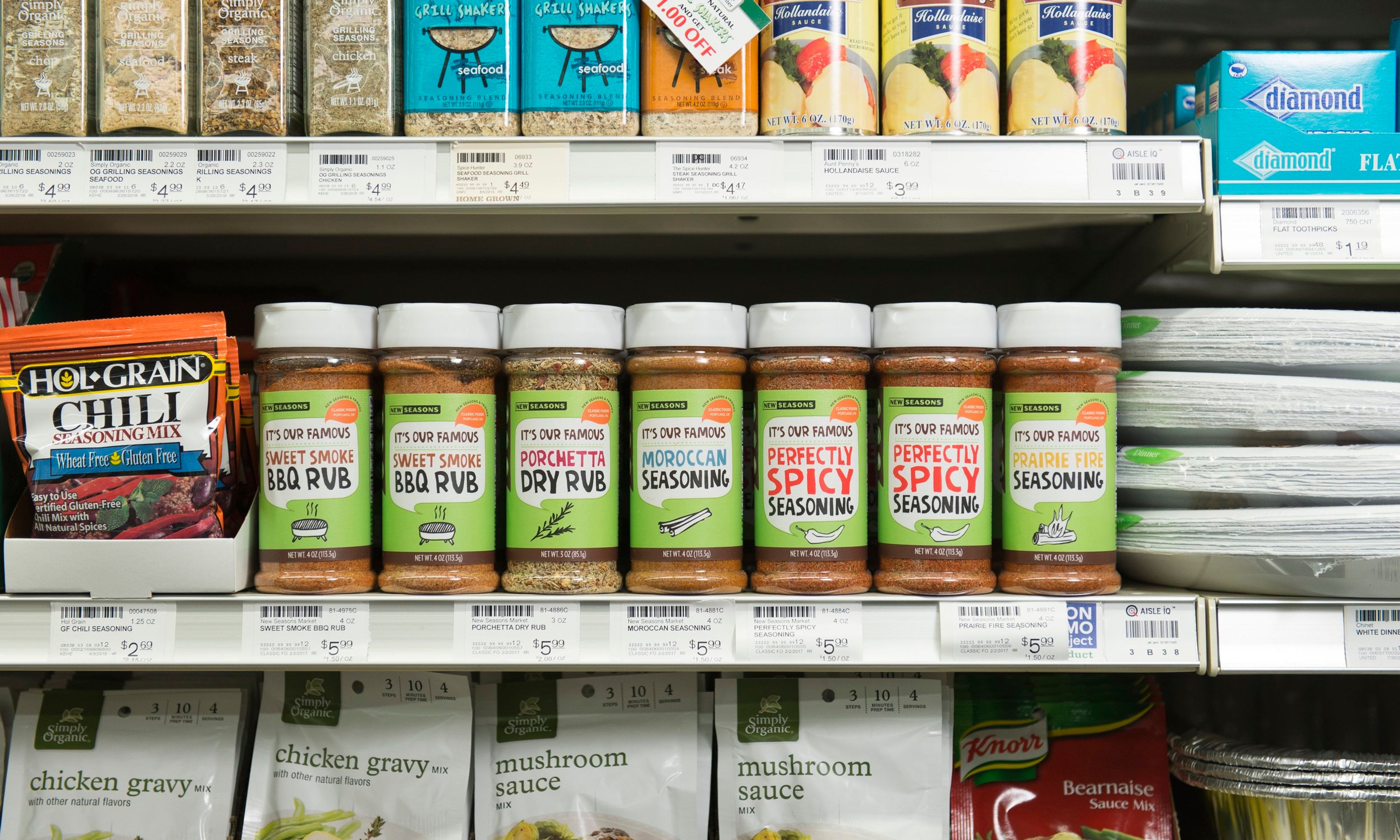 A row of seven spices made by New Seasons sitting on a grocery store shelf next to other spices, gravy, sauces, and paper plates