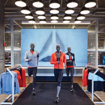 Three mannequins in running positions wearing sports attire in front of a large digital display