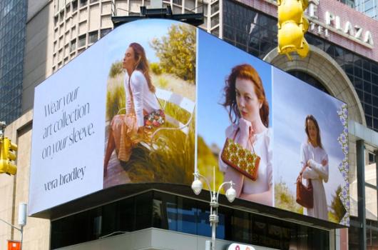 Billboard advertisement for Vera Bradley showcasing three women in outdoor settings, each featuring different handbags. The text on the left reads 'Wear your art collection on your sleeve.' The background highlights a sunny, natural environment, emphasizing the brand's focus on artistic and nature-inspired designs.