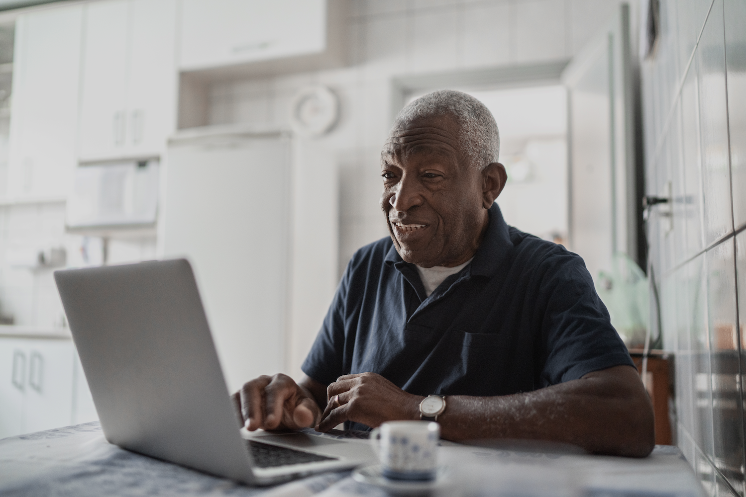 Elderly man sitting at a kitchen table, smiling and looking at an open laptop