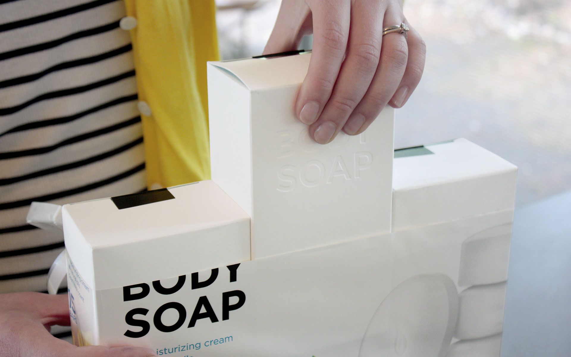 Person pulling a white rectangular box out of a pack of 3 that is wrapped in a thin plastic material that reads “Body Soap”