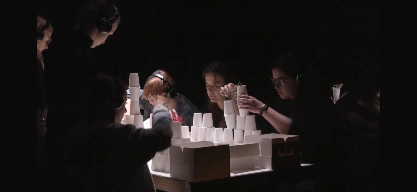 Público con auriculares alrededor de una mesa iluminada, construyendo una torre de vasos