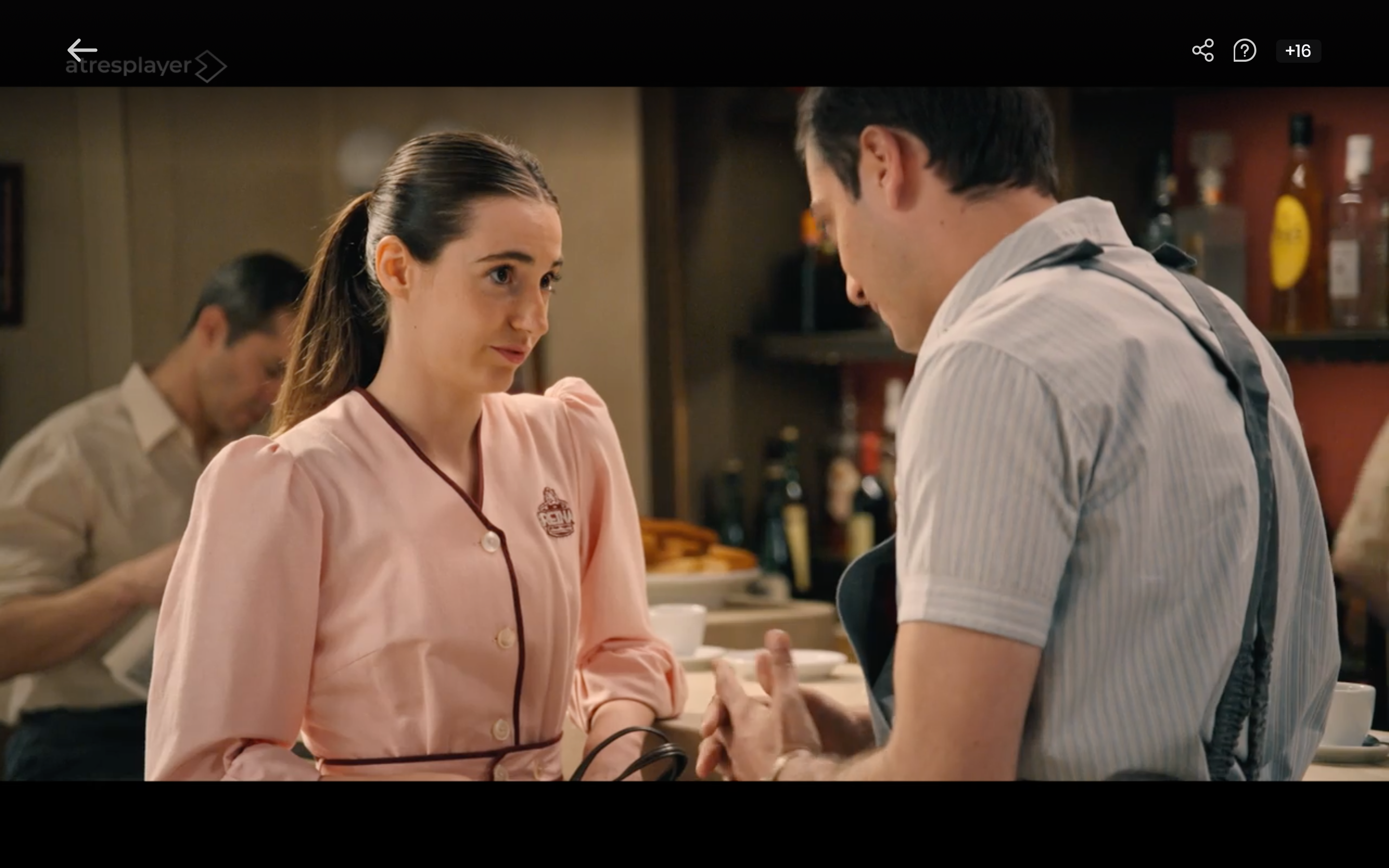 Cecilia Mansilla como camarera, de uniforme rosa, hablando con un compañero en la barra de un bar