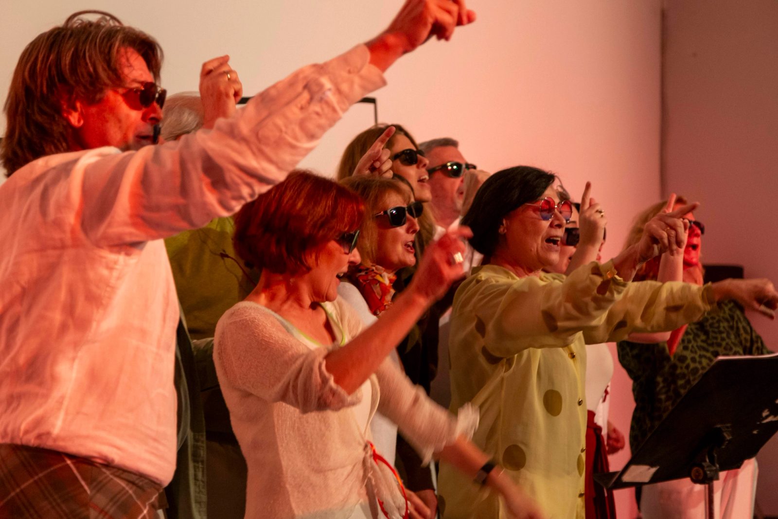 Grupo de participantes con gafas de sol cantando y señalando al frente, bajo una luz rosada