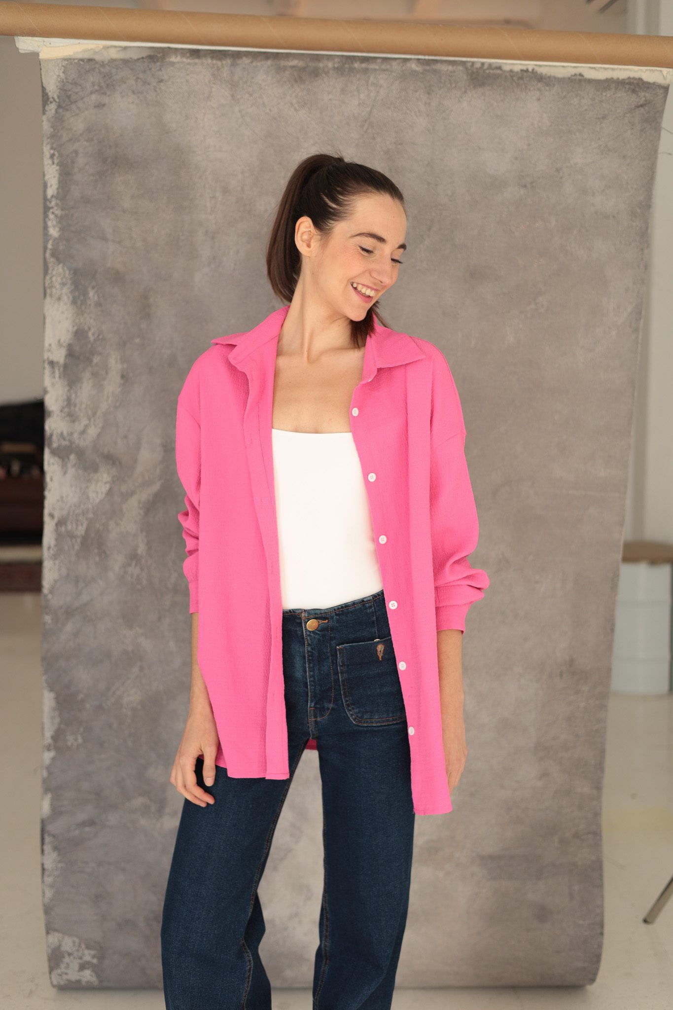 Cecilia Mansilla de cuerpo entero, con camisa rosa y vaqueros, sonriendo sobre un fondo gris de estudio