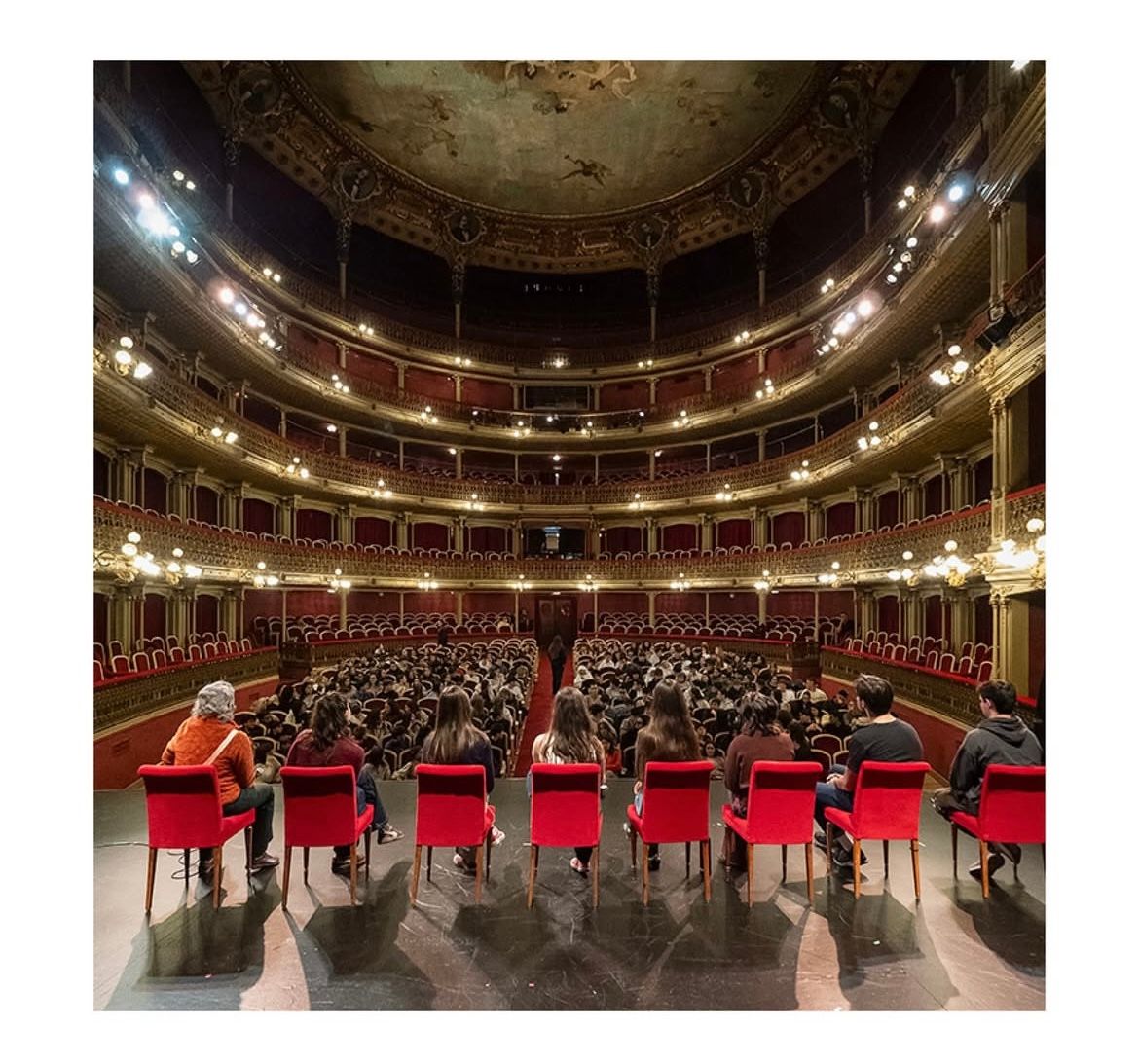 Un grupo sentado en el escenario de un teatro a la italiana, de espaldas al patio de butacas lleno de público