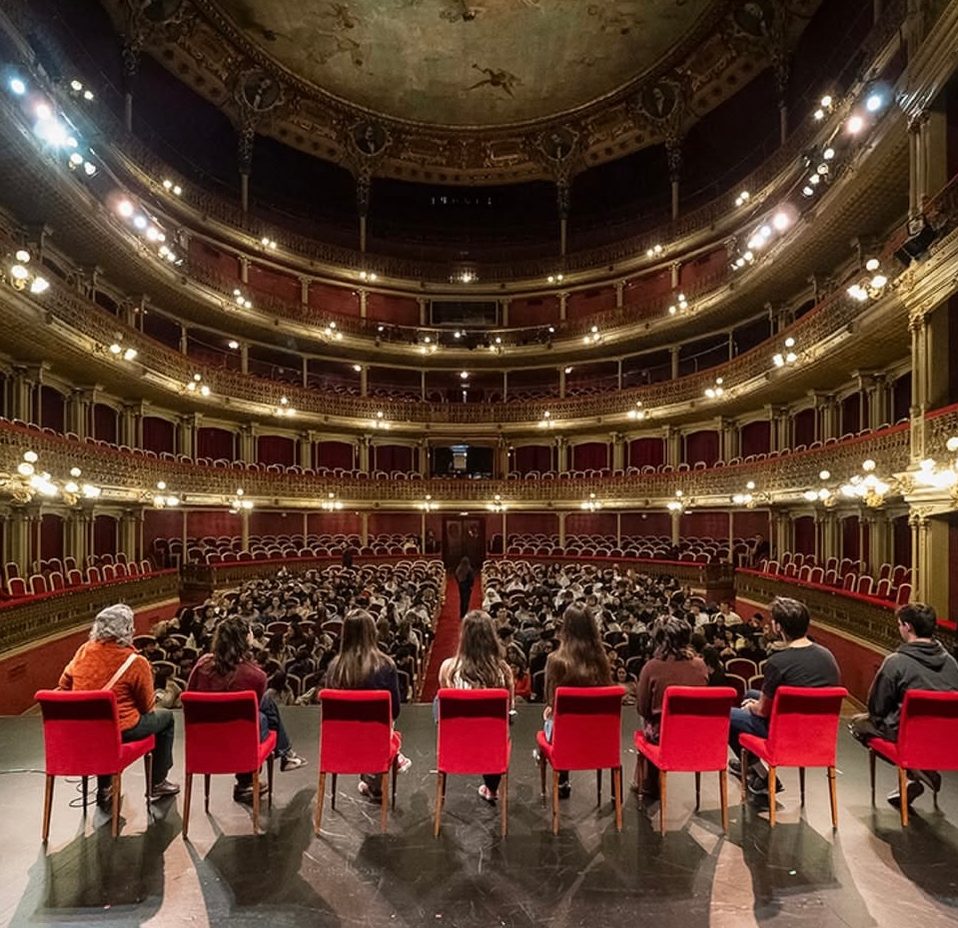 Un grupo sentado en el escenario de un teatro a la italiana, de espaldas al patio de butacas lleno de público