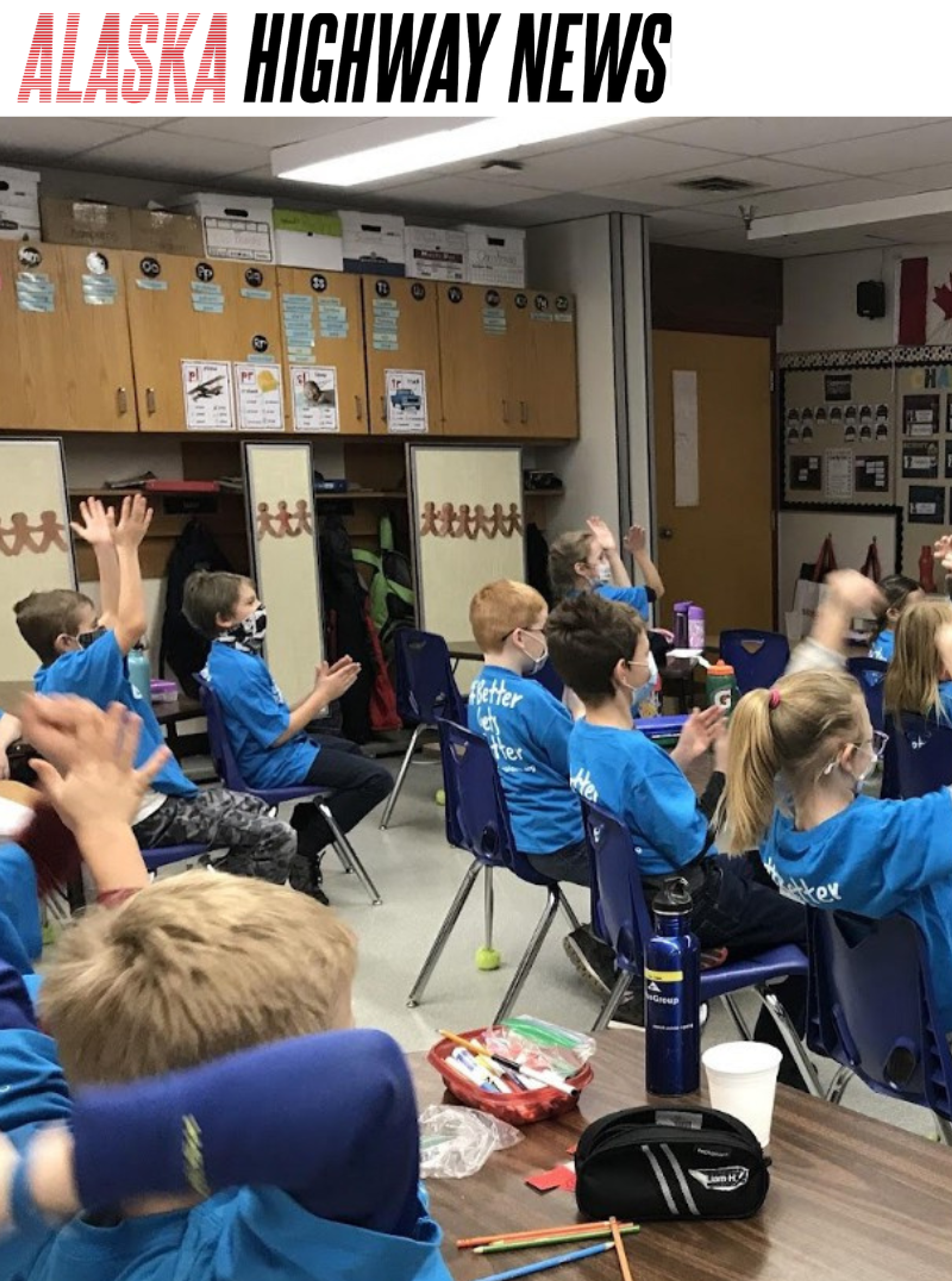 Students in matching blue T-shirts raise their hands and clap during a classroom mentoring session, under an Alaska Highway News banner.