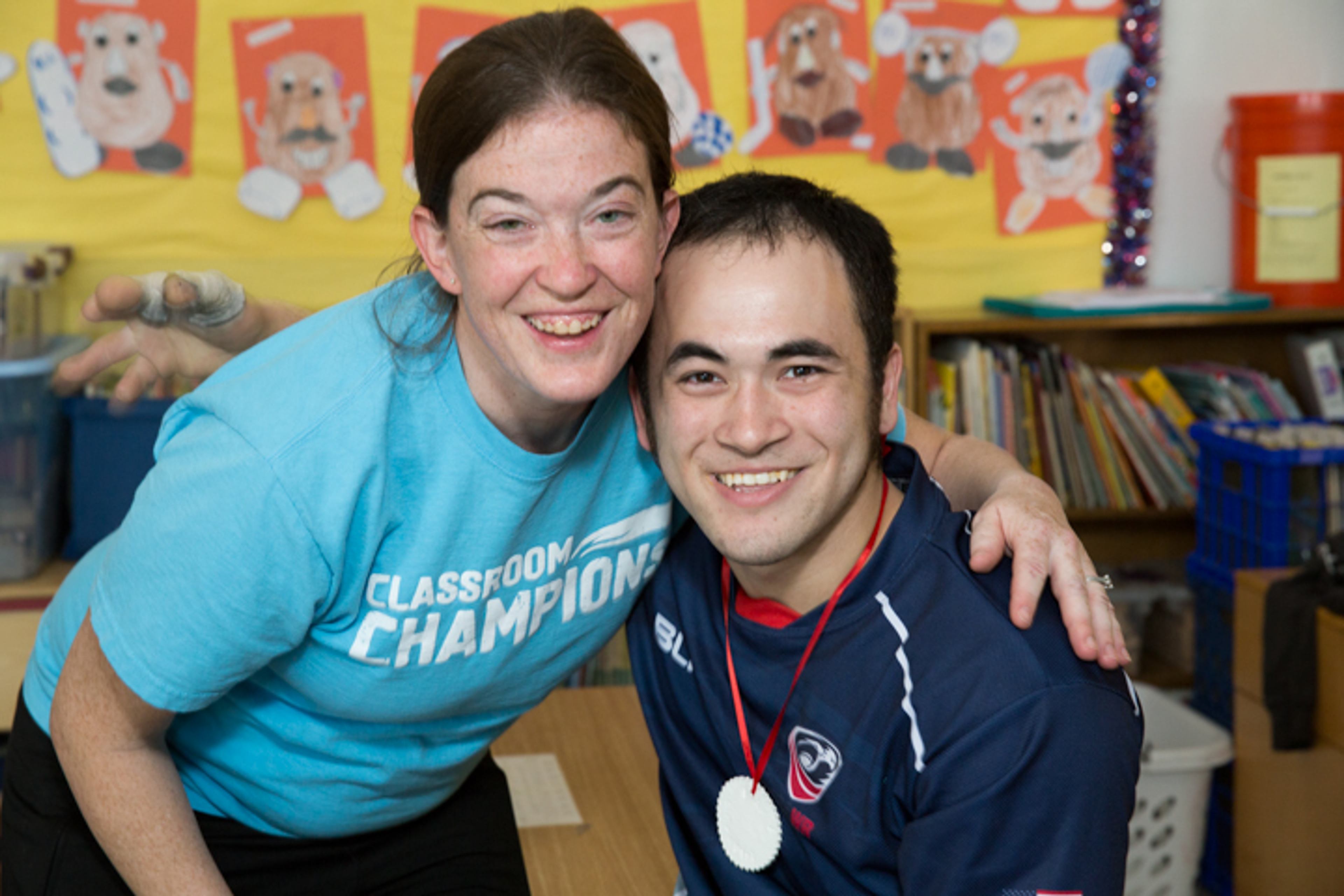 A Classroom Champions mentor wearing a medal poses with a volunteer in a classroom.
