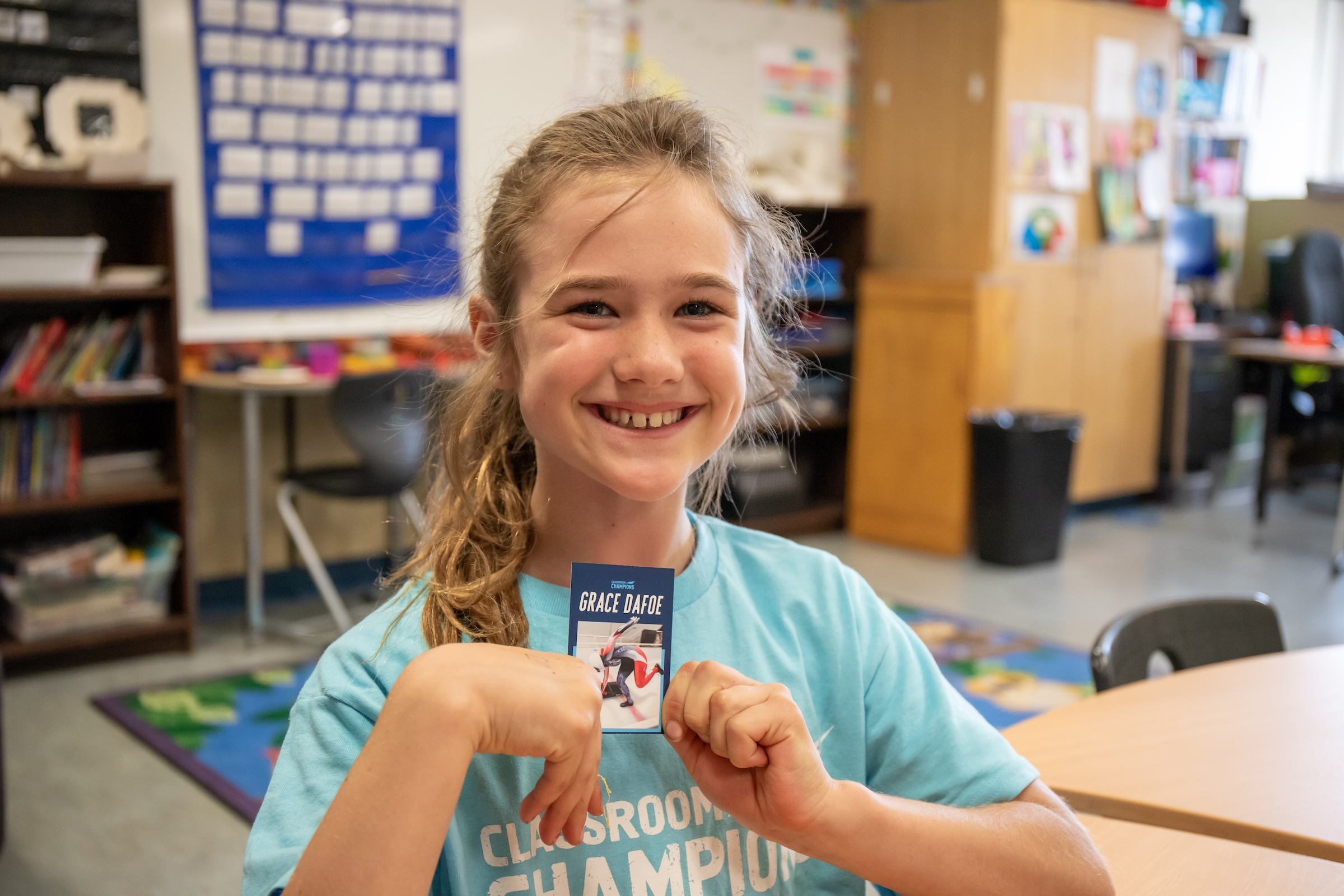 A smiling student in a Classroom Champions shirt holds up a trading card of mentor Grace Dafoe.