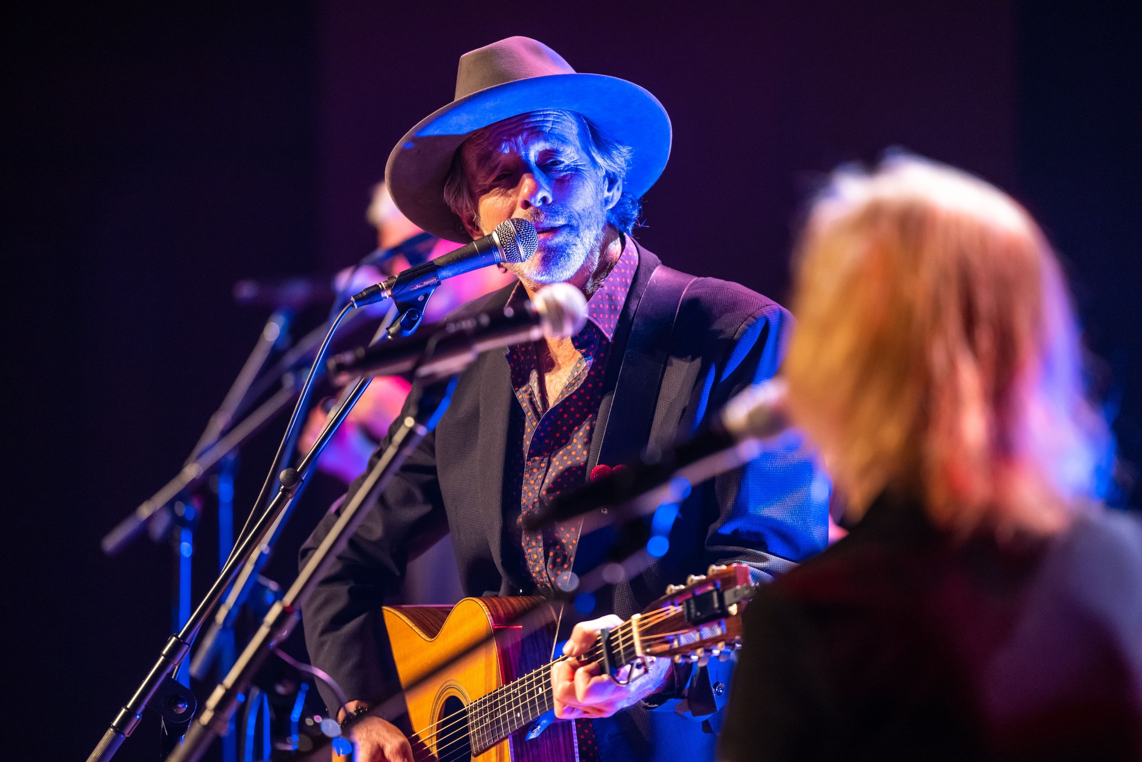 A musician wearing a hat sings and plays acoustic guitar on stage at a Concert for Classrooms benefit.