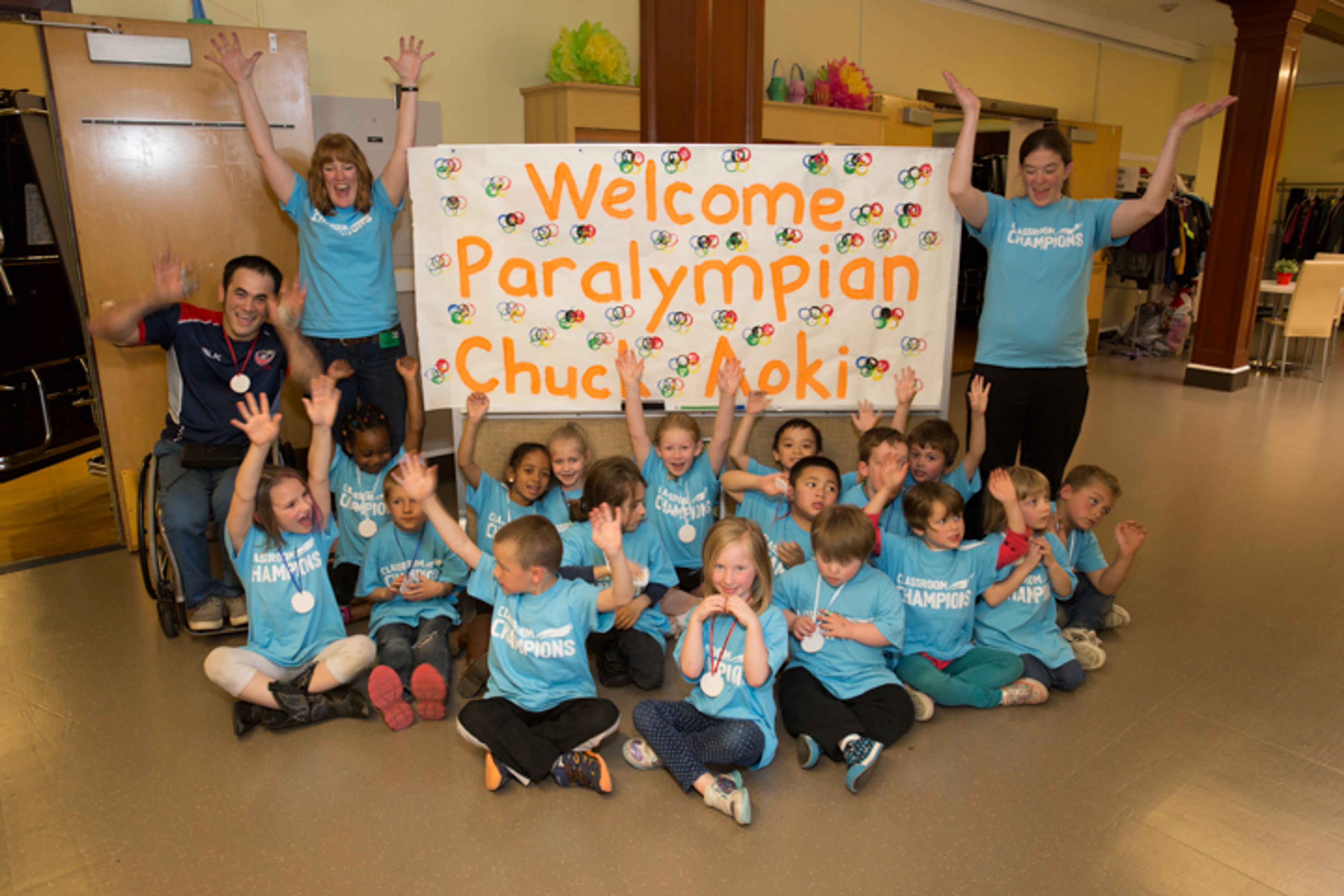 Paralympian Chuck Aoki and teachers cheer with a classroom of students beneath a welcome banner.