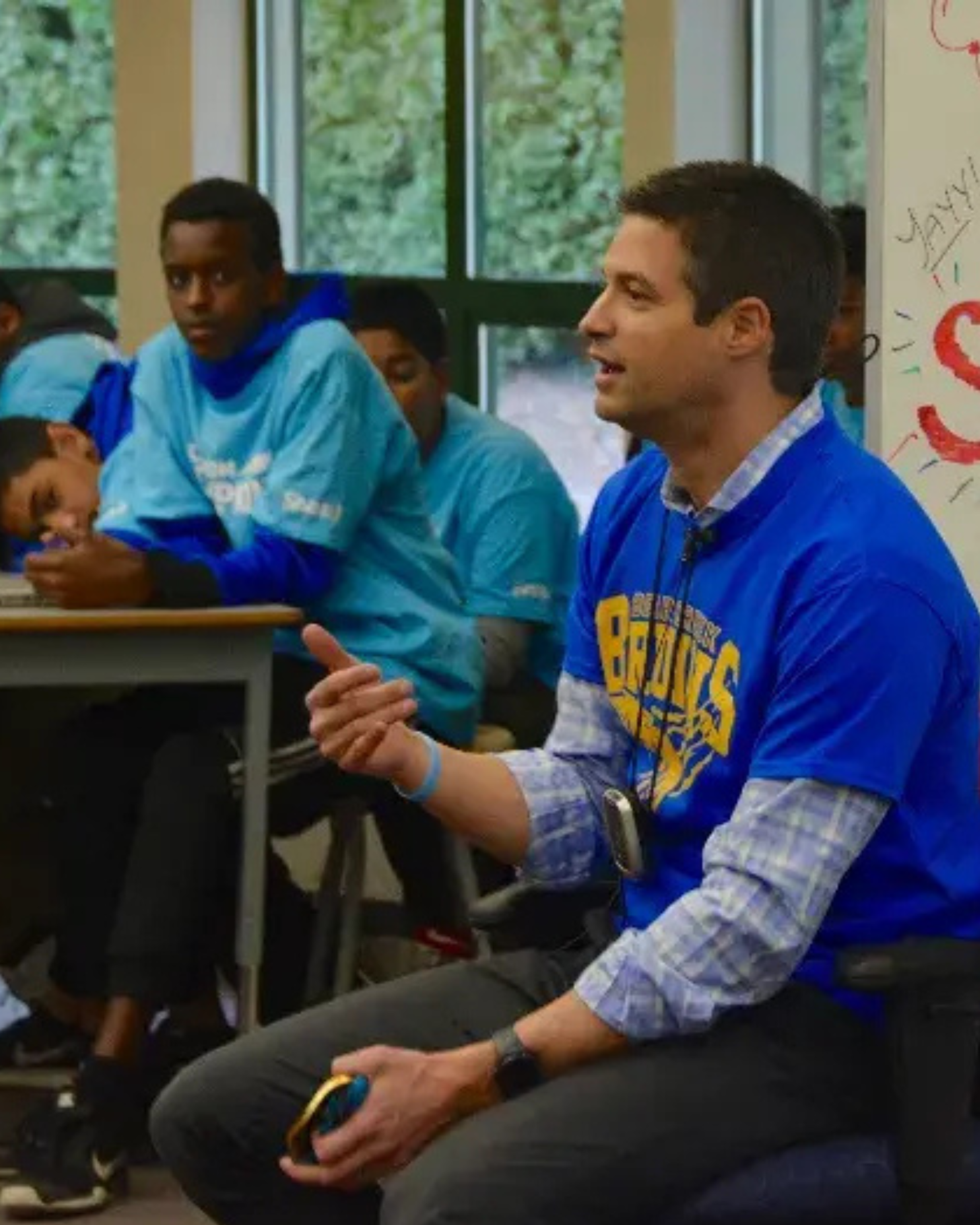 An athlete mentor speaks with students gathered around desks in a classroom.