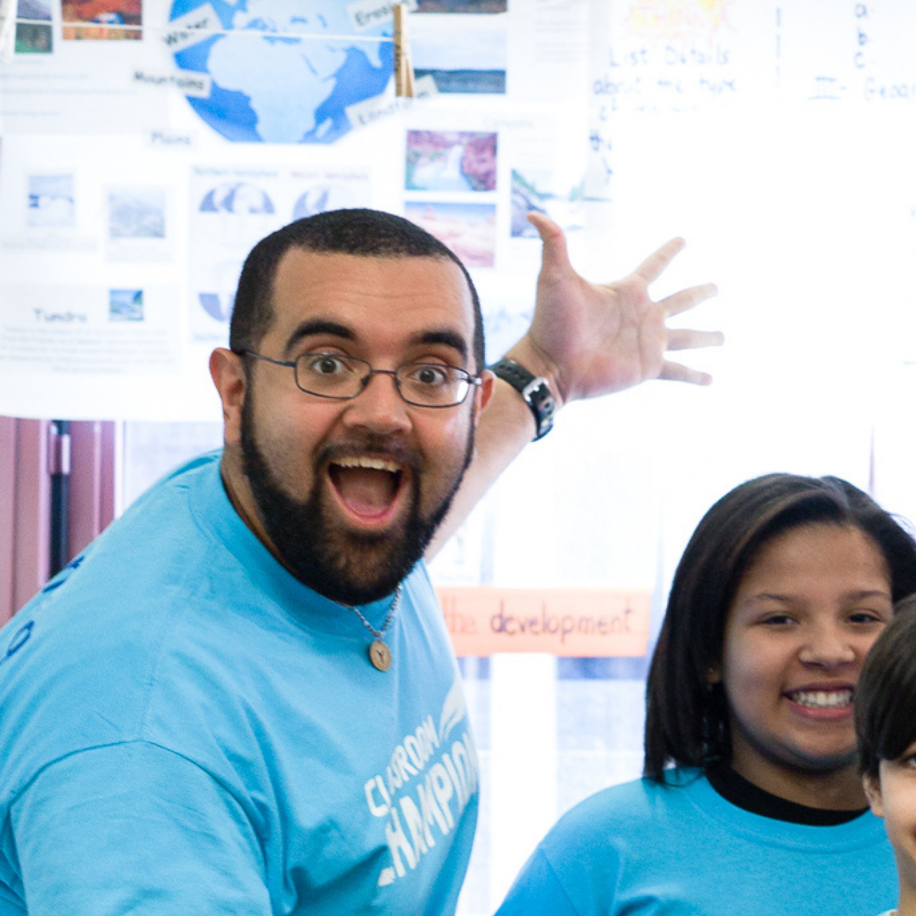 A teacher and students smile enthusiastically in a classroom decorated with geography posters.