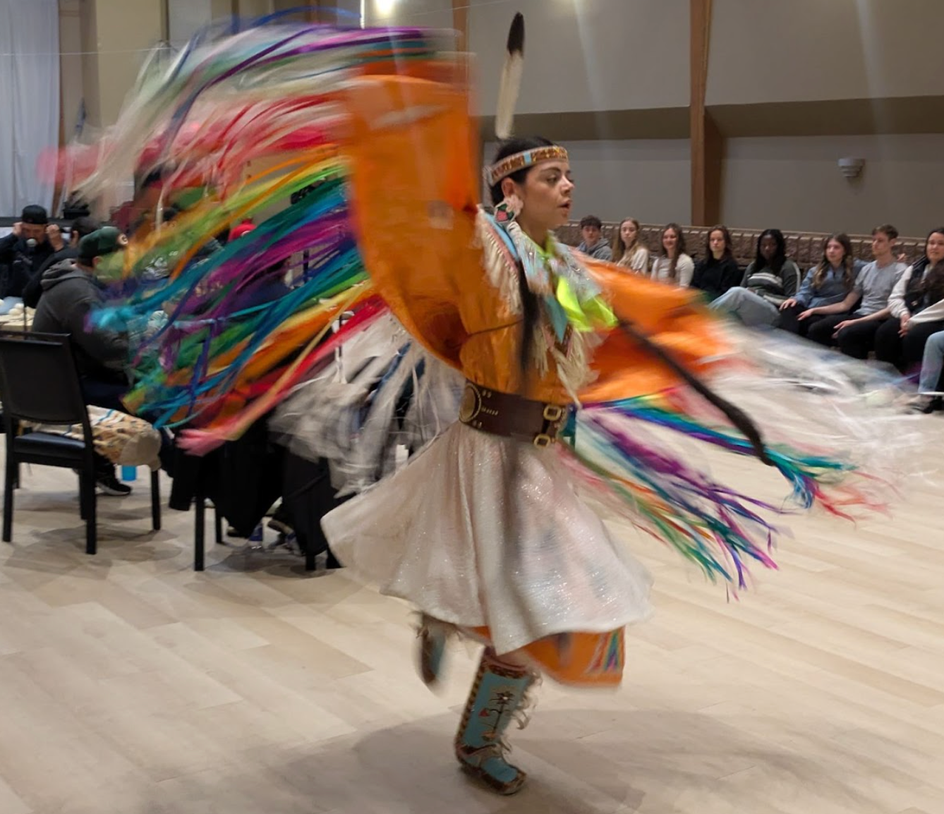A jingle dress dancer in colorful ribbon regalia performs for a seated audience during a traditional dance.