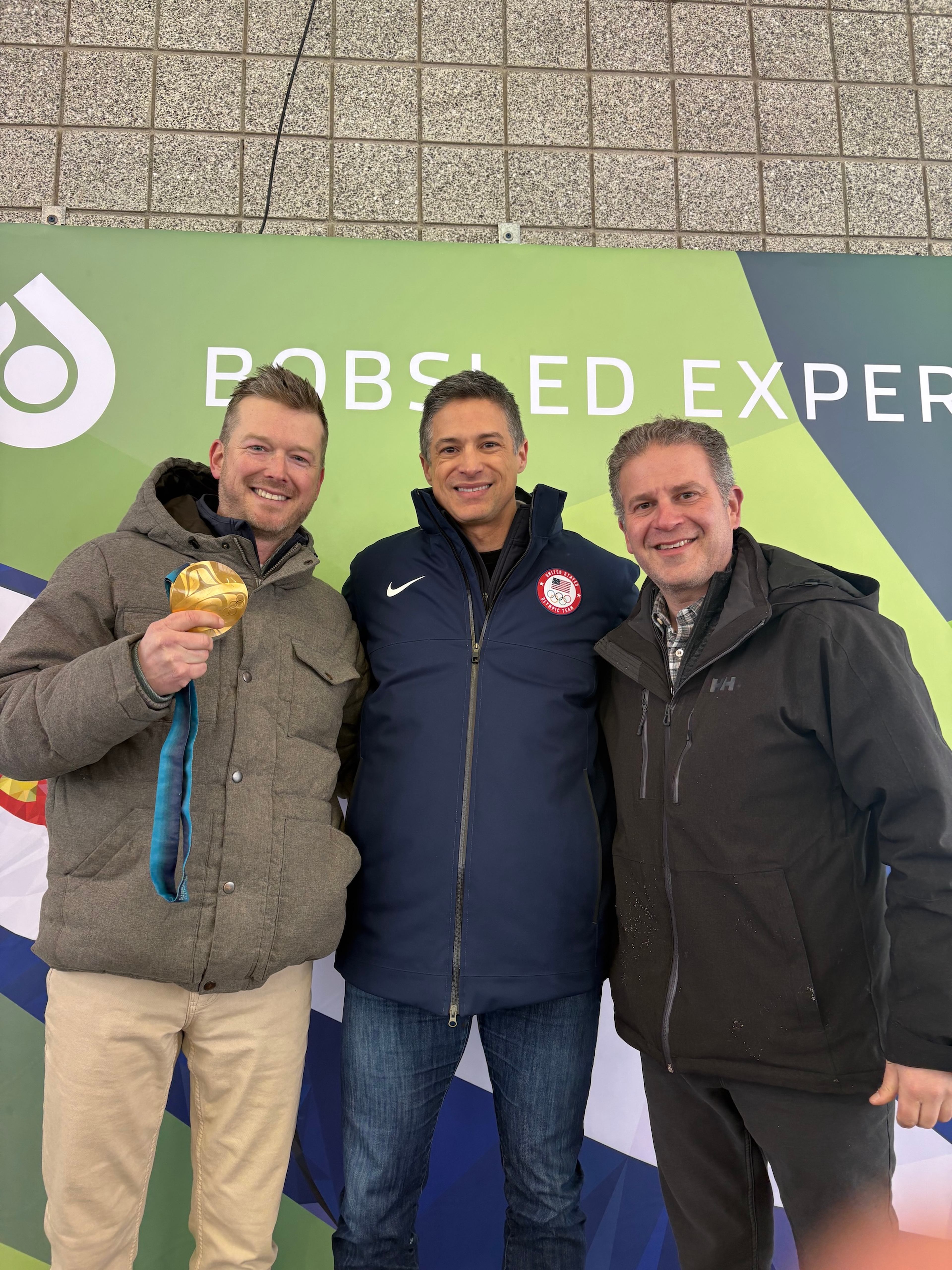 A mentor holds up his Olympic gold medal while posing with two men at a Bobsled Experience event.