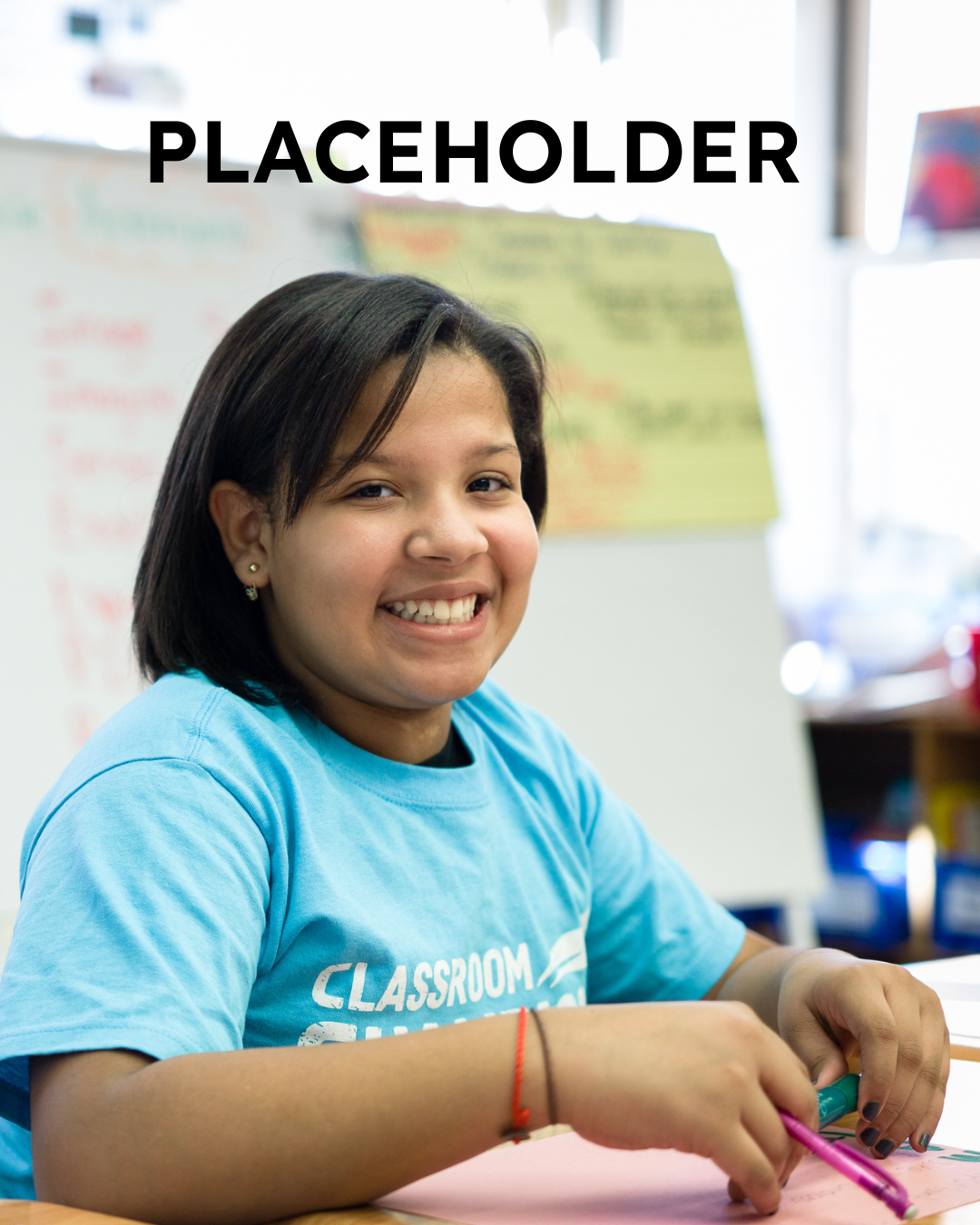 A smiling student wearing a Classroom Champions T-shirt sits at a desk working on schoolwork.