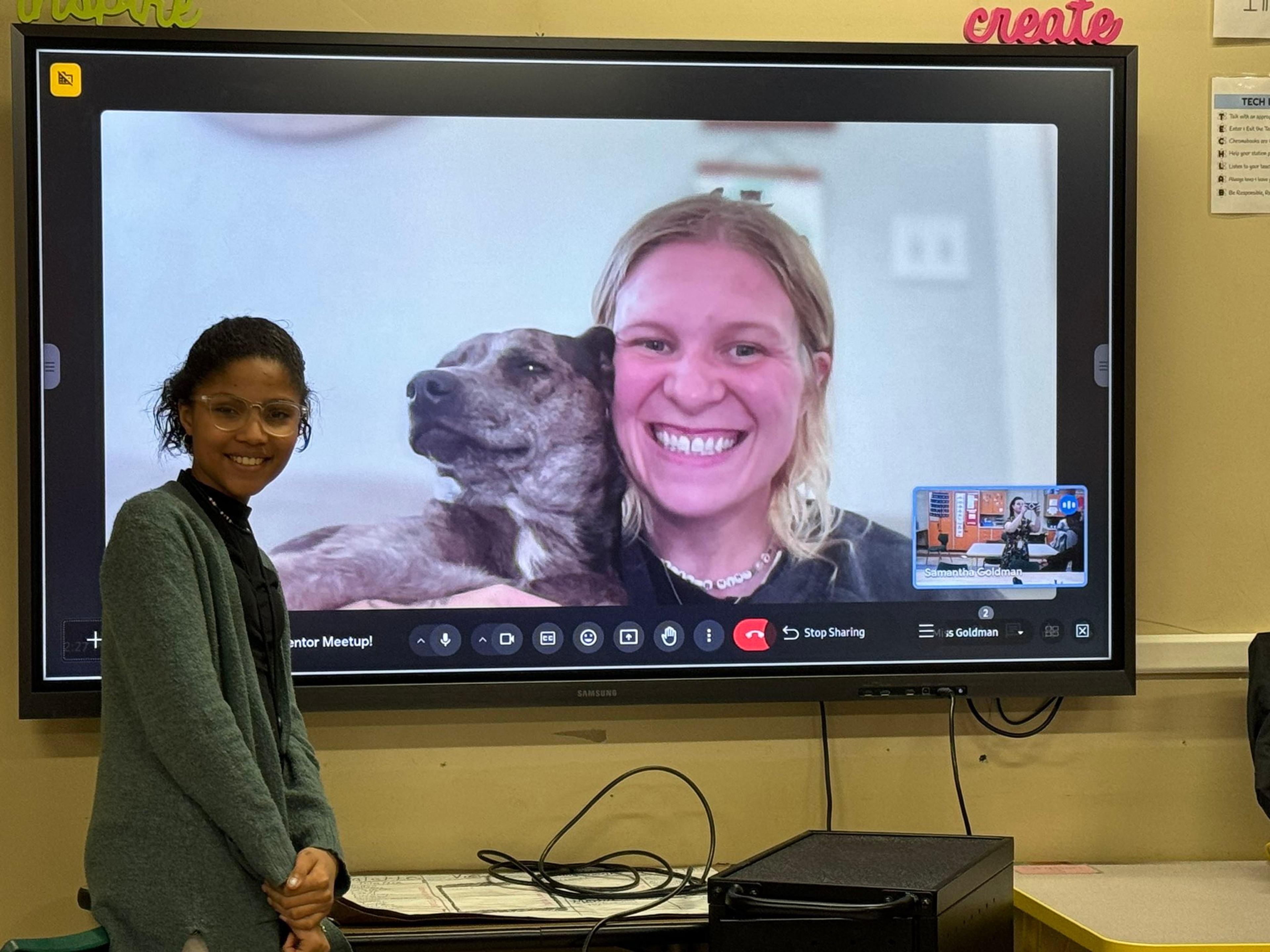 A student stands beside a classroom screen during a video call with her athlete mentor and dog.