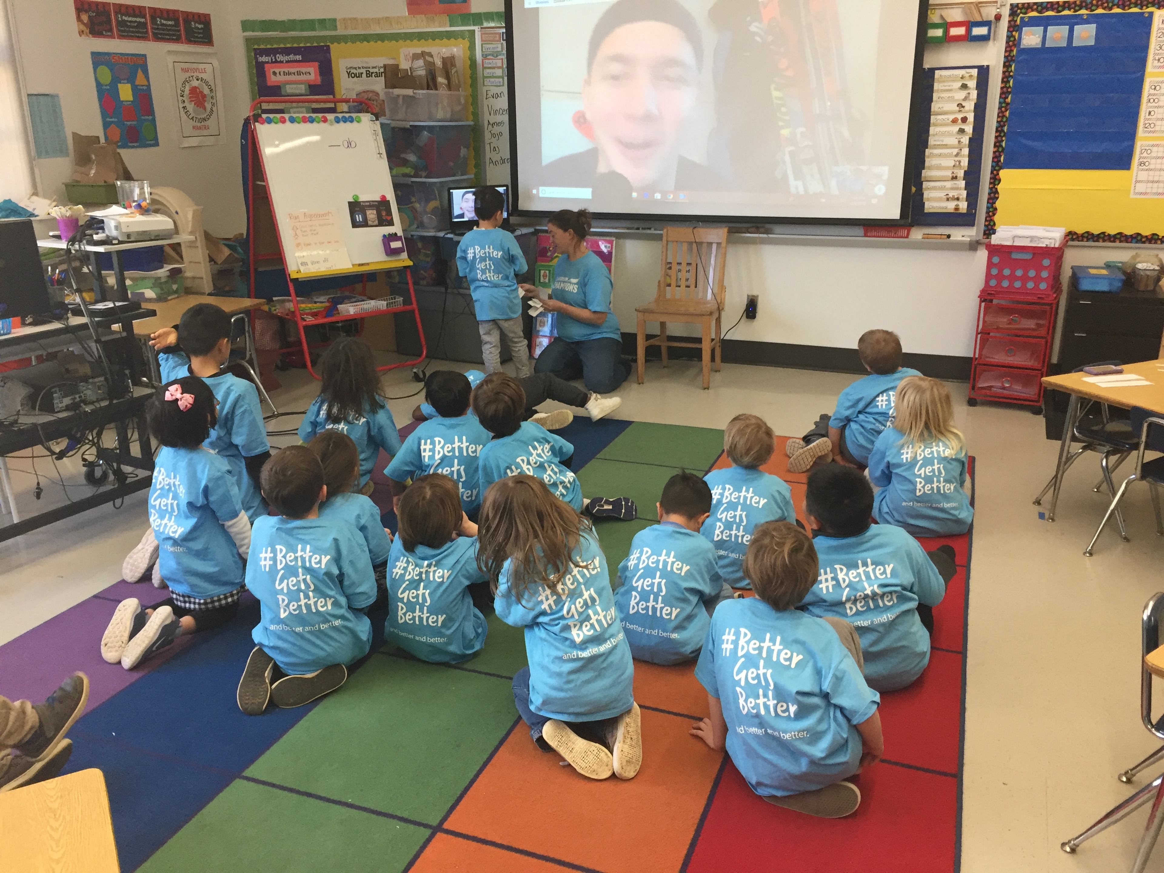 Students wearing "#BetterGetsBetter" shirts sit on a classroom rug during a video call with an athlete mentor.