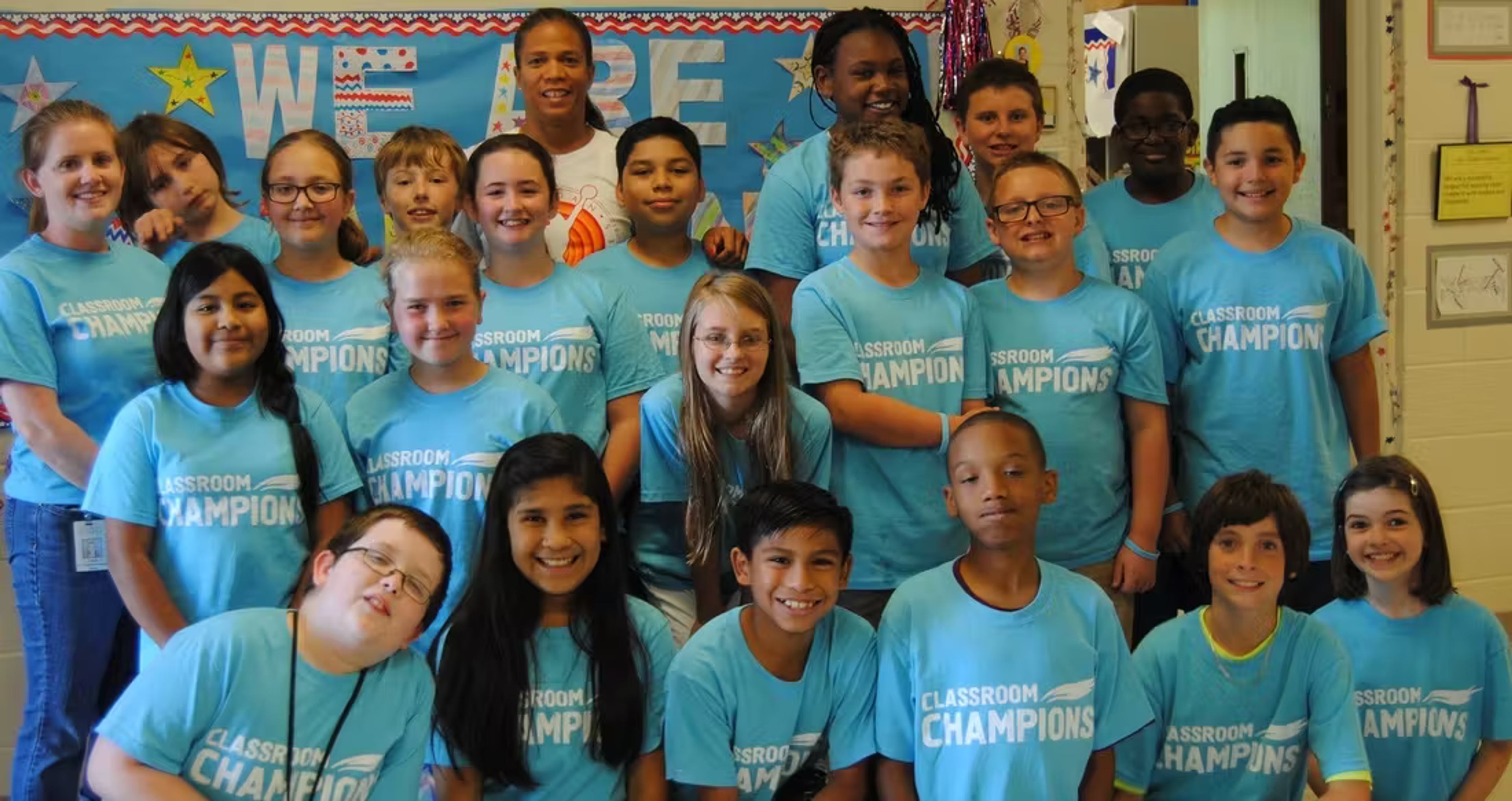 A classroom of students and their teacher pose together wearing matching Classroom Champions T-shirts.