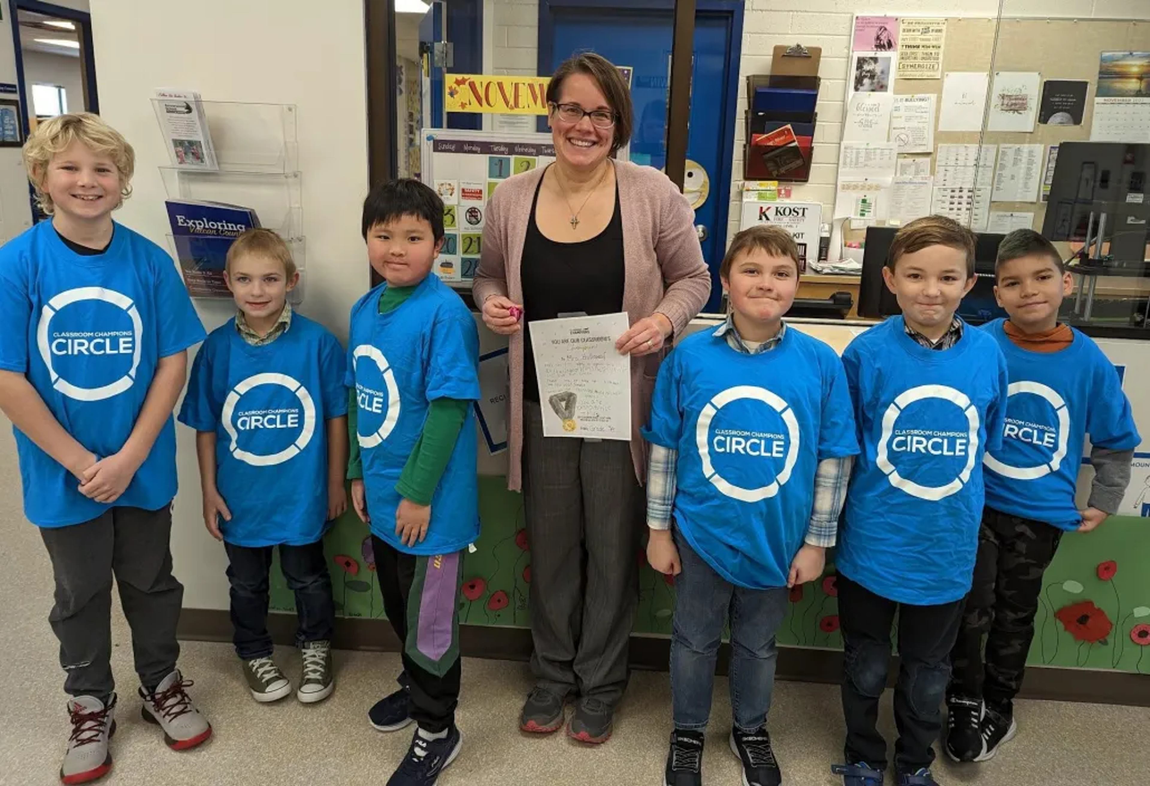 A teacher holds a Classroom Champions Circle award certificate alongside students wearing matching program t-shirts.