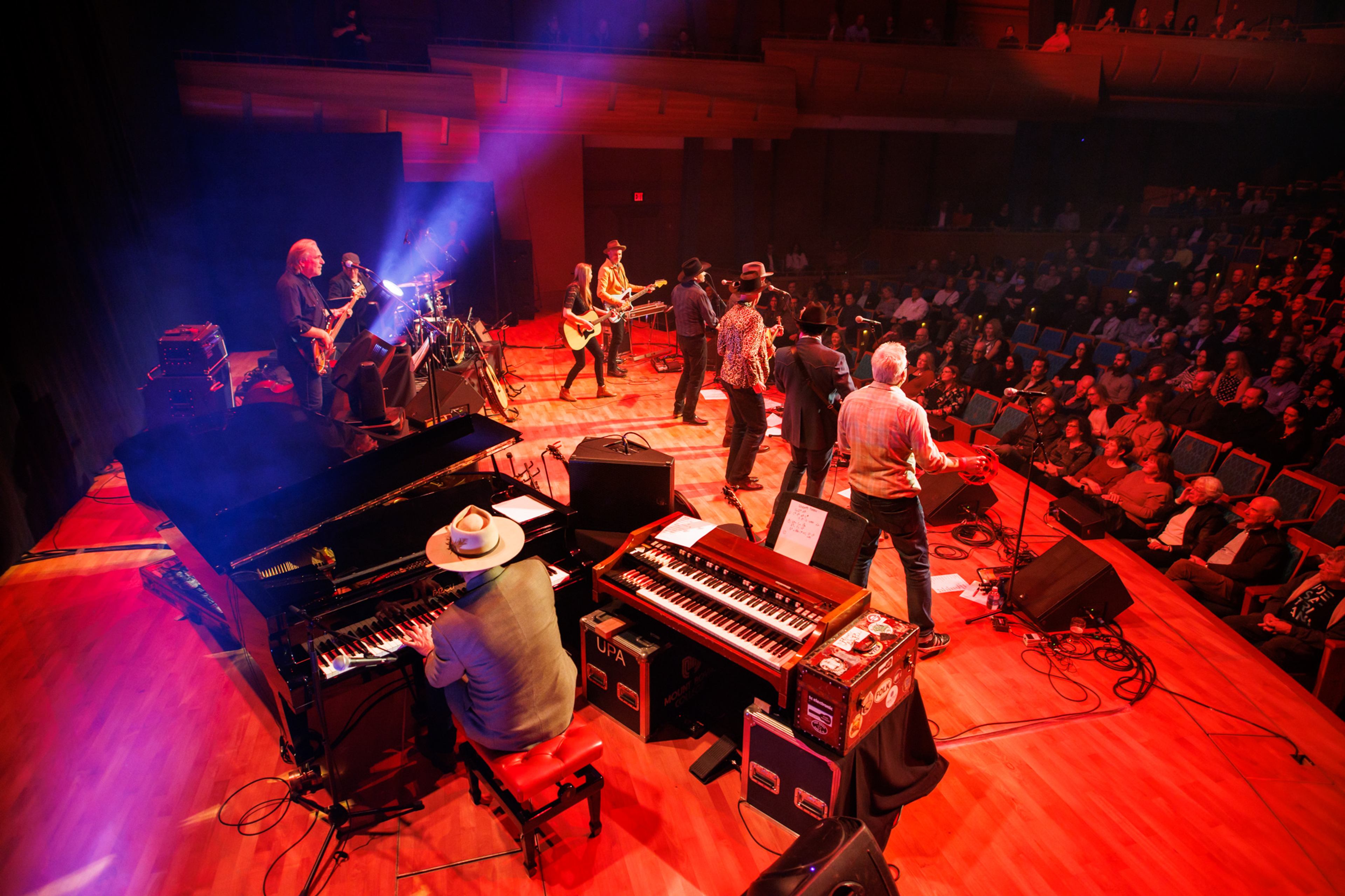 A band performs on stage with piano and organ before a full theater audience at a Concert for Classrooms event.