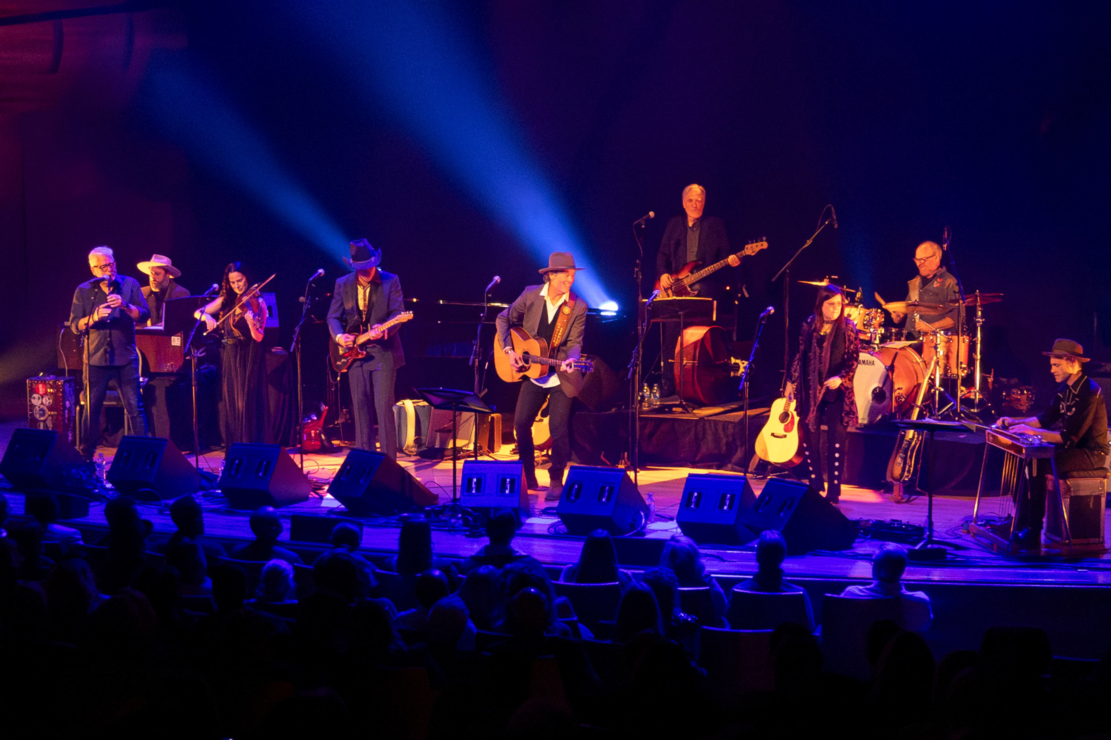 A full band performs on stage under blue lights at the Concert for Classrooms Calgary benefit show.