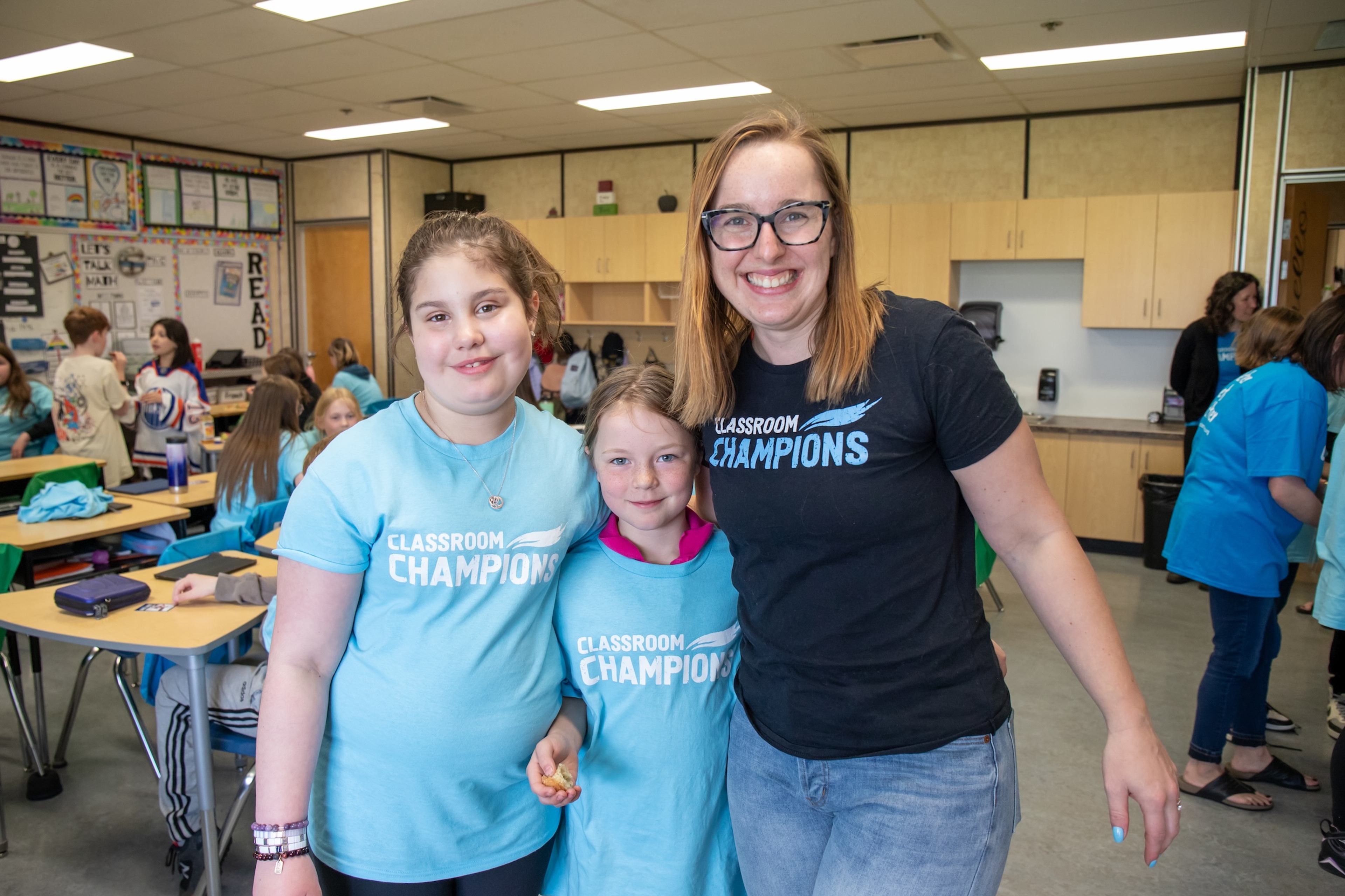 A teacher and two students smile together wearing Classroom Champions t-shirts in their classroom.
