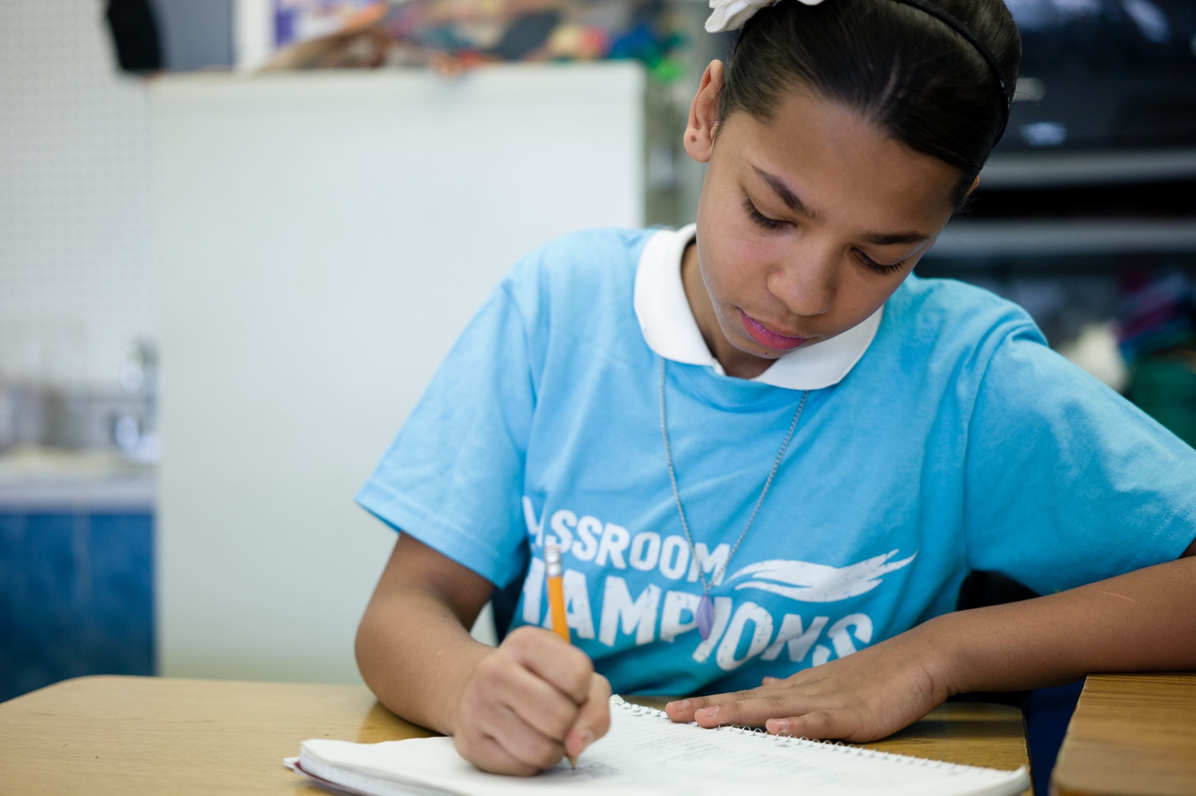 A student wearing a Classroom Champions t-shirt writes in a notebook at her desk.
