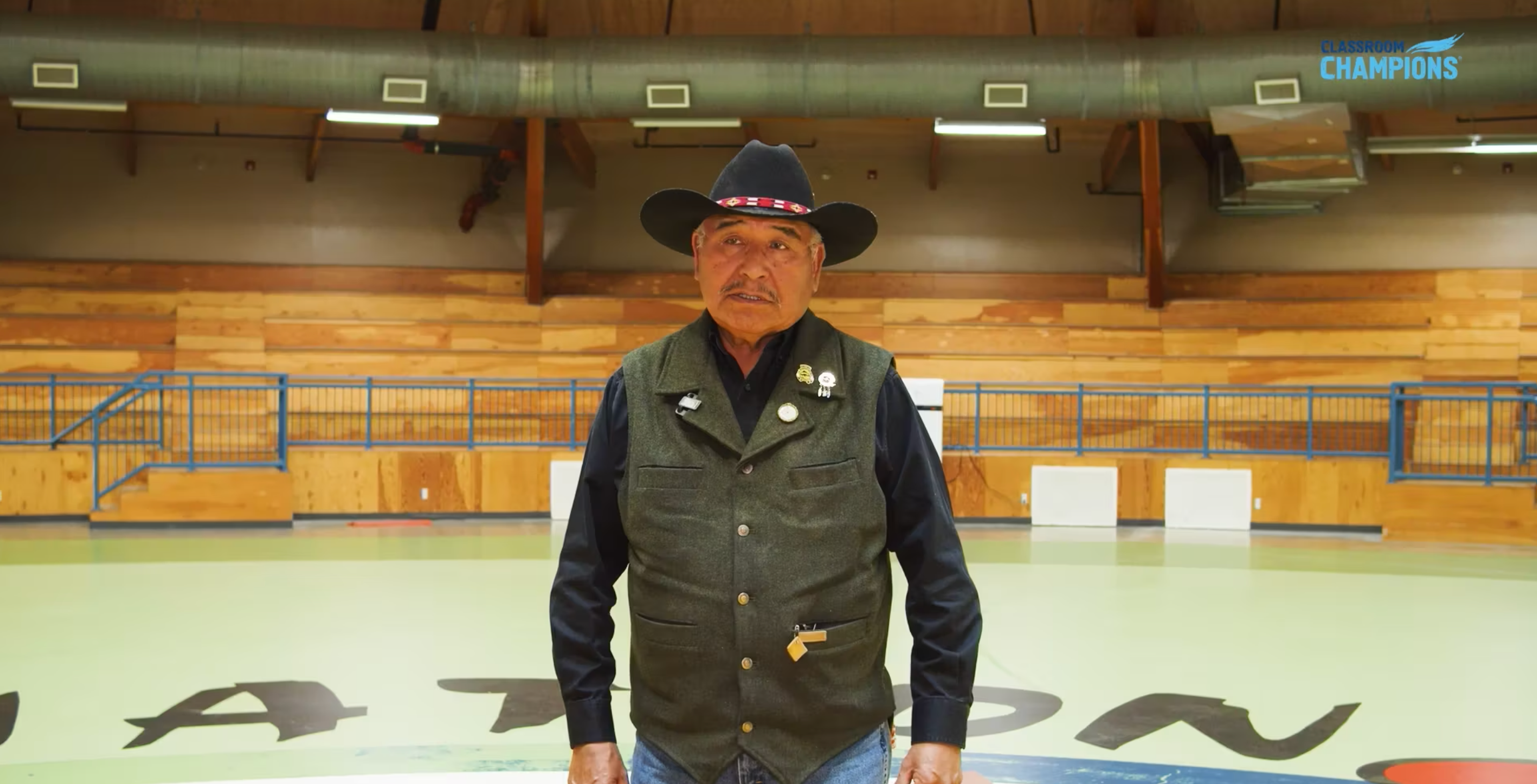 A man wearing a hat and vest stands in a gymnasium, speaking as part of the Indigenous Circle Program.