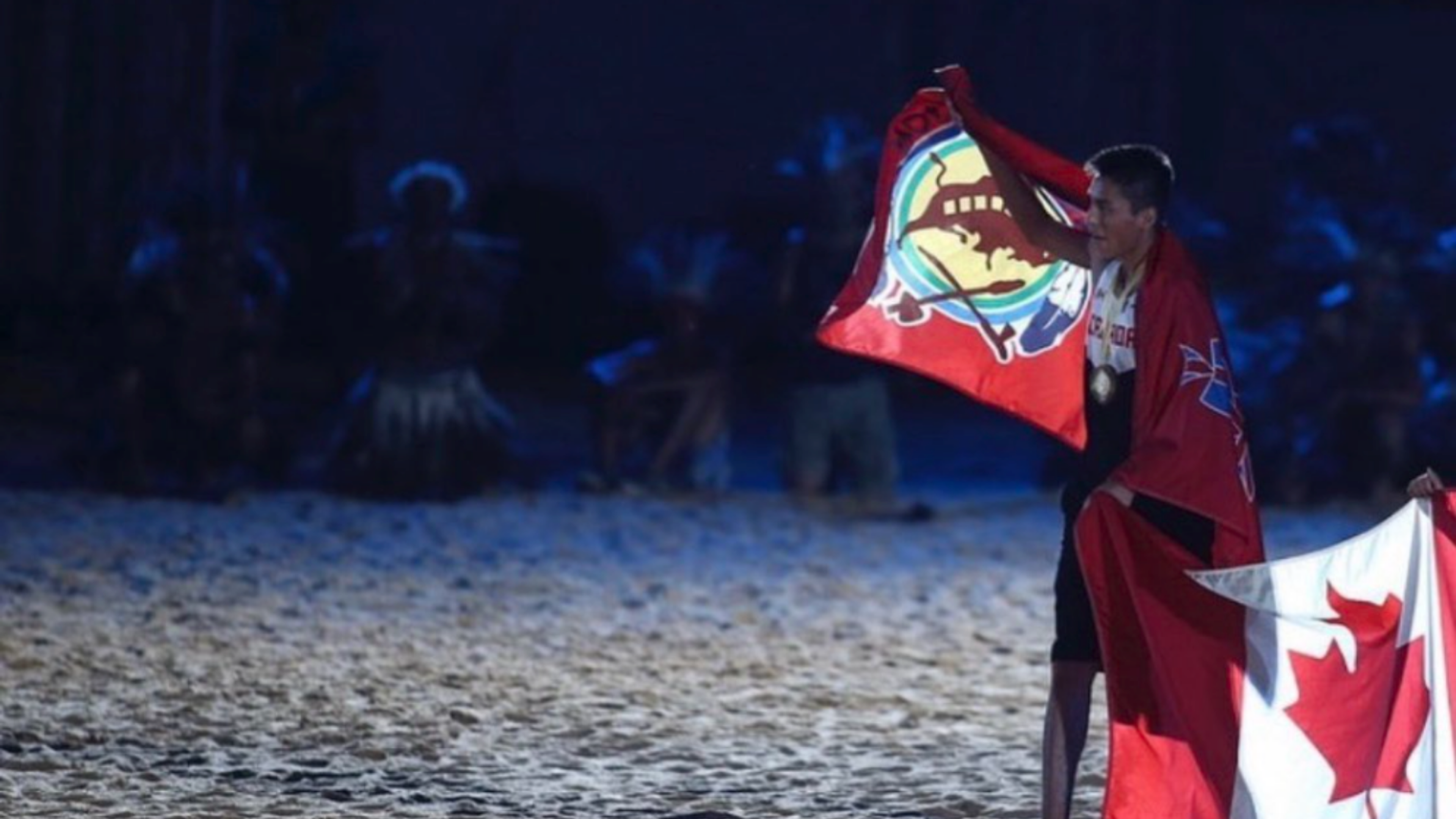An athlete wearing a medal waves an Indigenous flag alongside a Canadian flag at a nighttime ceremony.