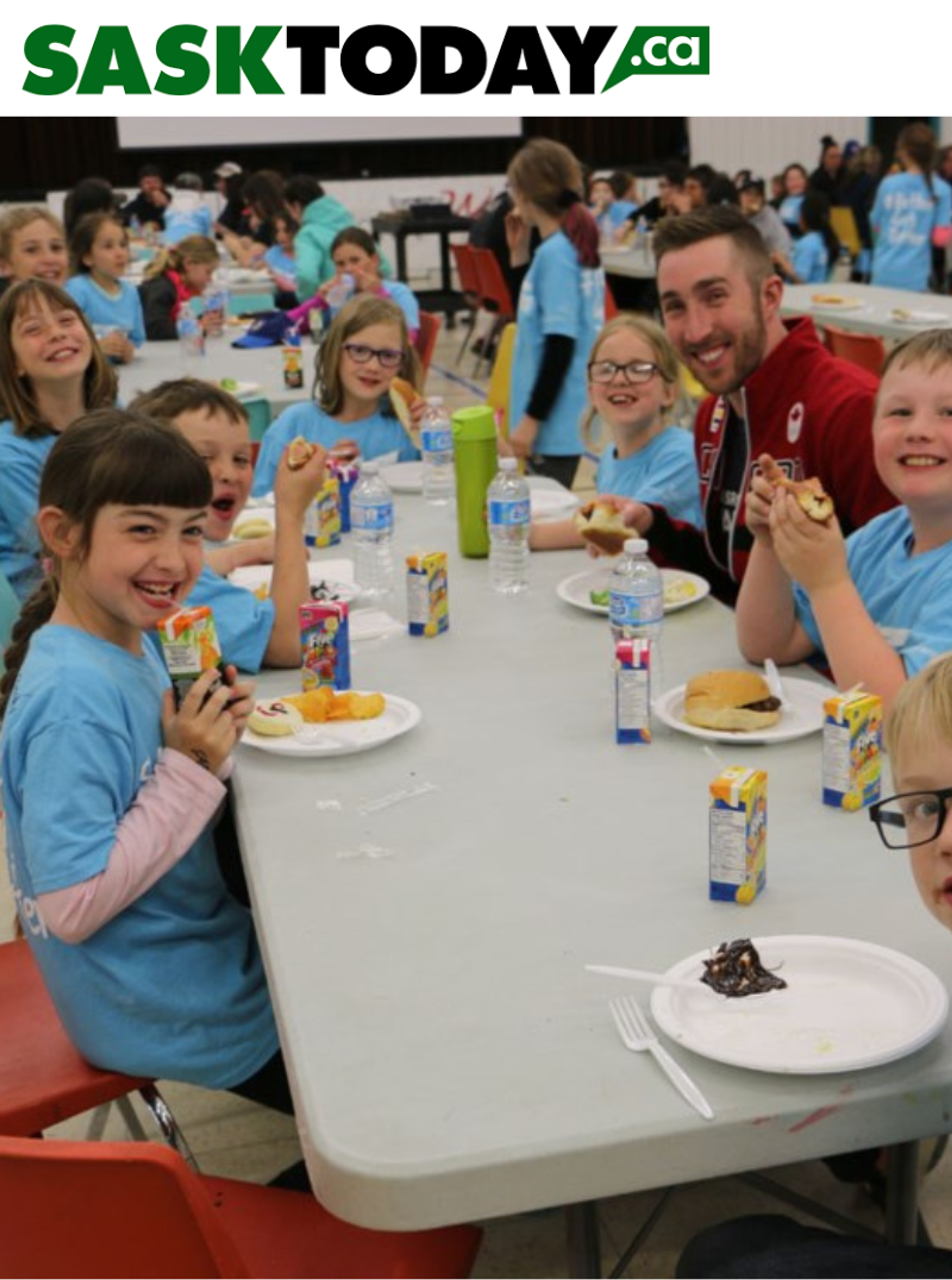 An athlete mentor shares lunch and smiles with students wearing matching blue T-shirts at a school event.
