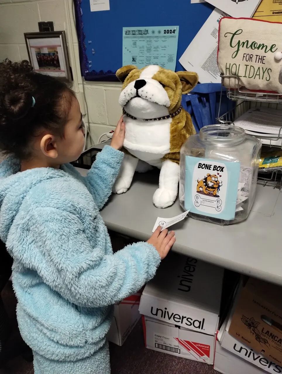 A student places a note into a classroom "Bone Box" beside a stuffed bulldog mascot.