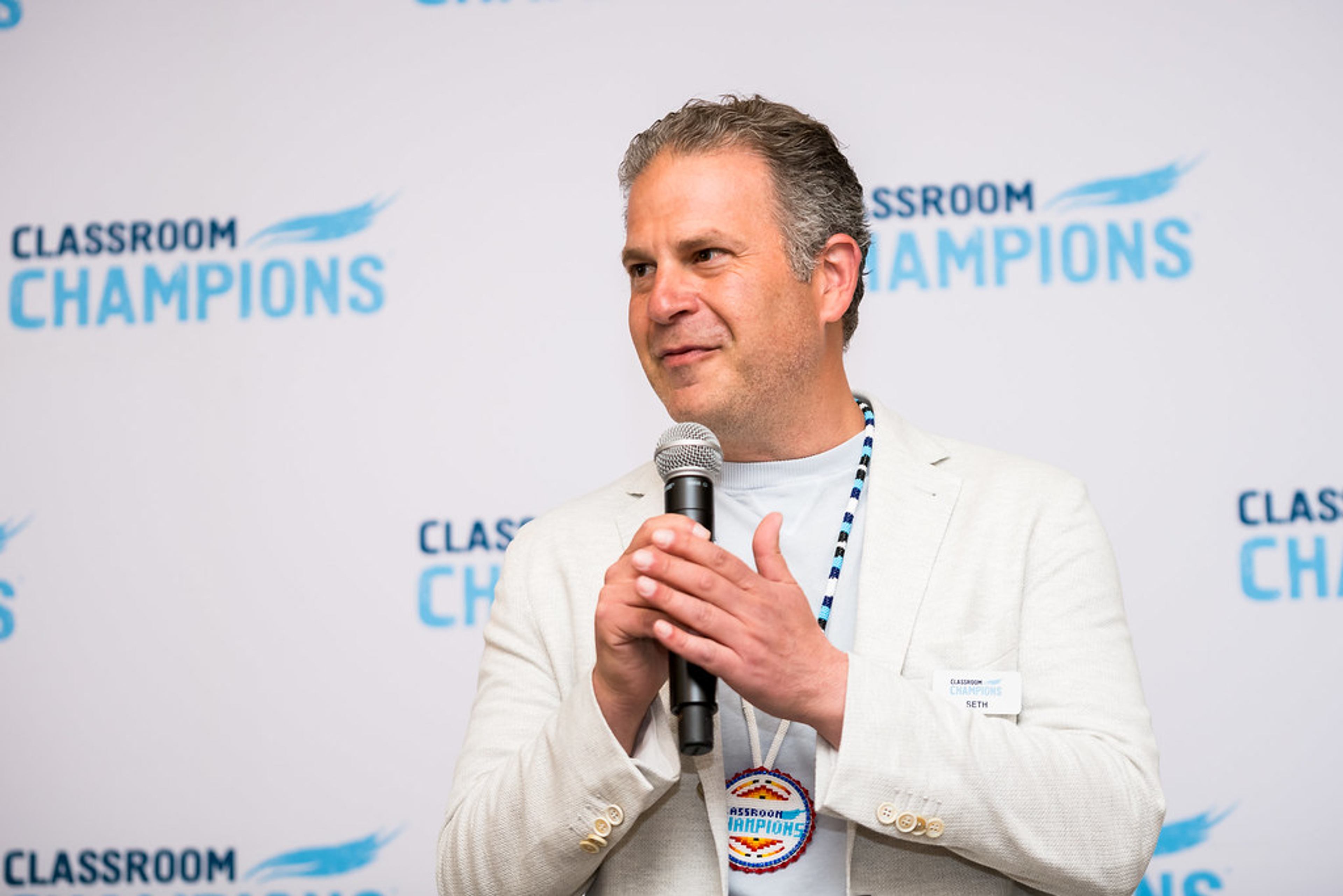 A speaker addresses the audience with a microphone in front of a Classroom Champions backdrop.