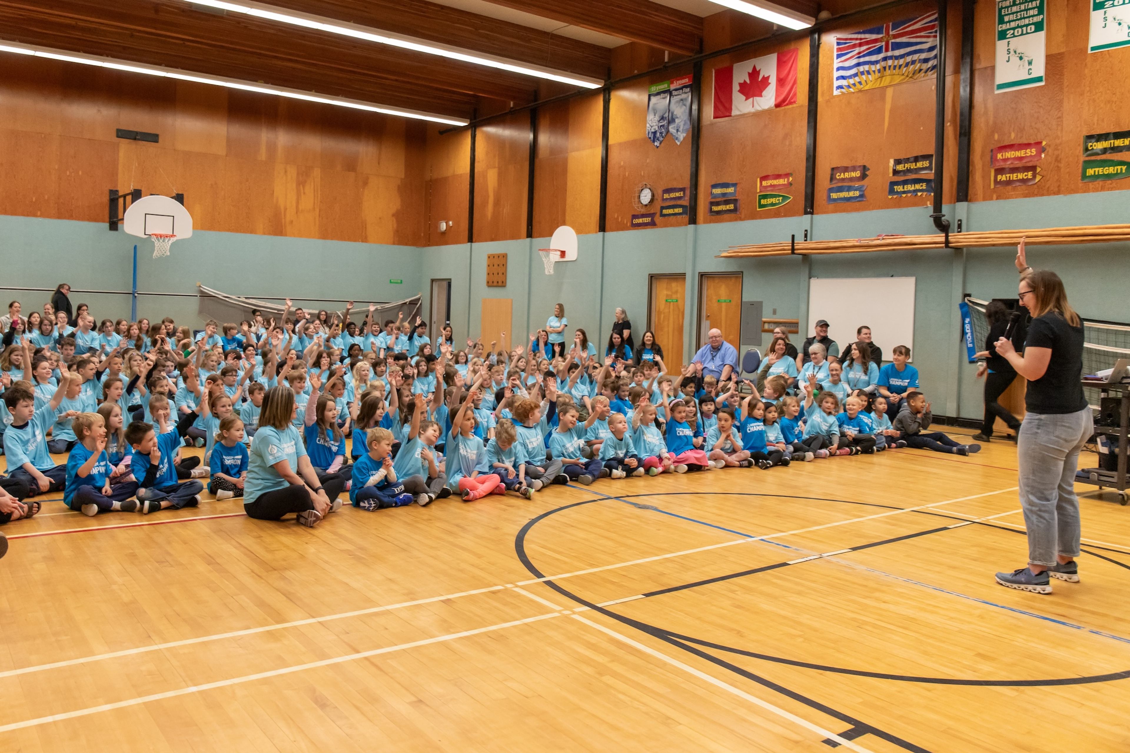Students in Classroom Champions T-shirts sit in a gym, raising their hands as a speaker addresses the assembly.